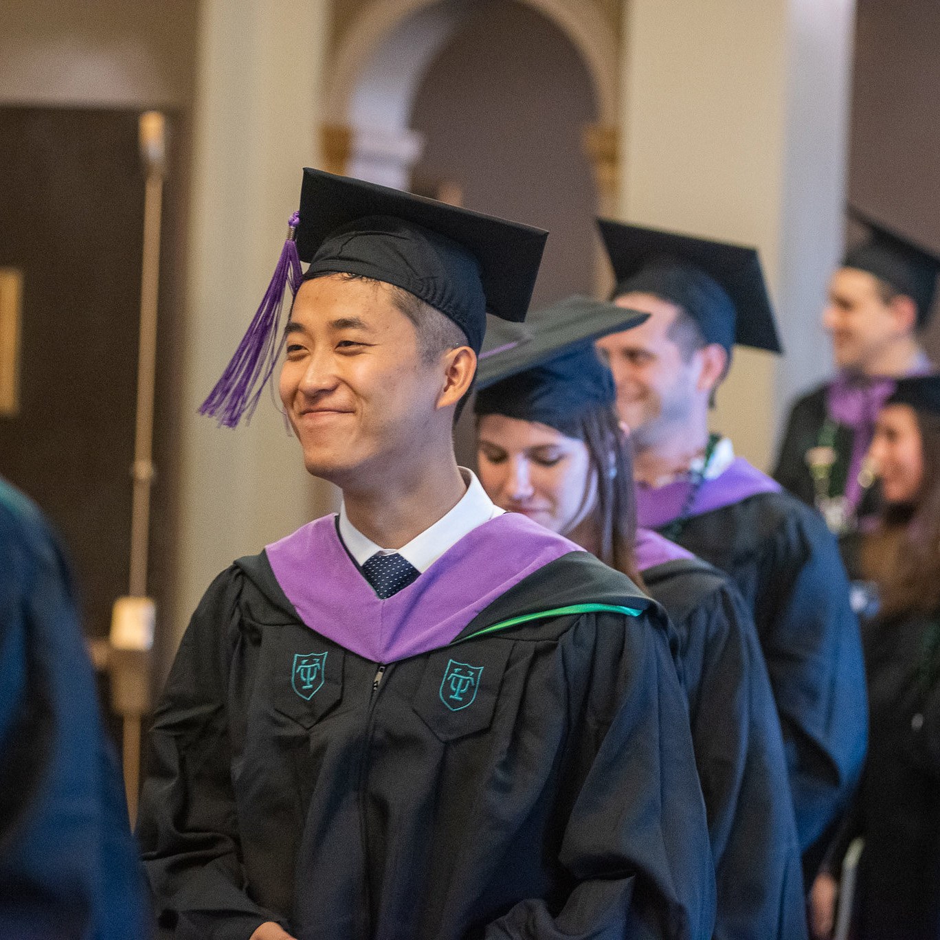 Student smiling in graduation cap and gown with line of fellow graduates behind him.