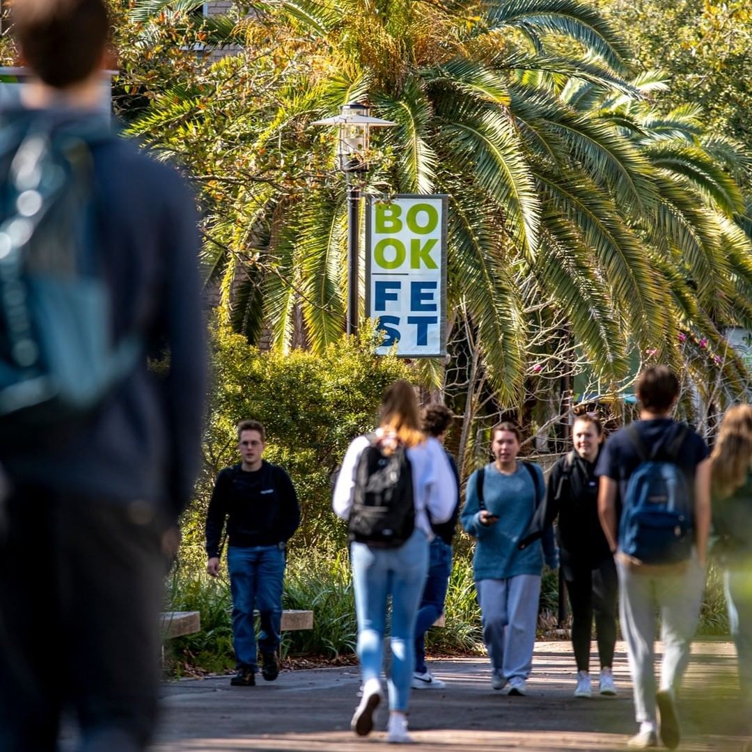 People walk outside on a sidewalk with palm trees overhead and in the background is a streetlight pole banner that reads "Book Fest"