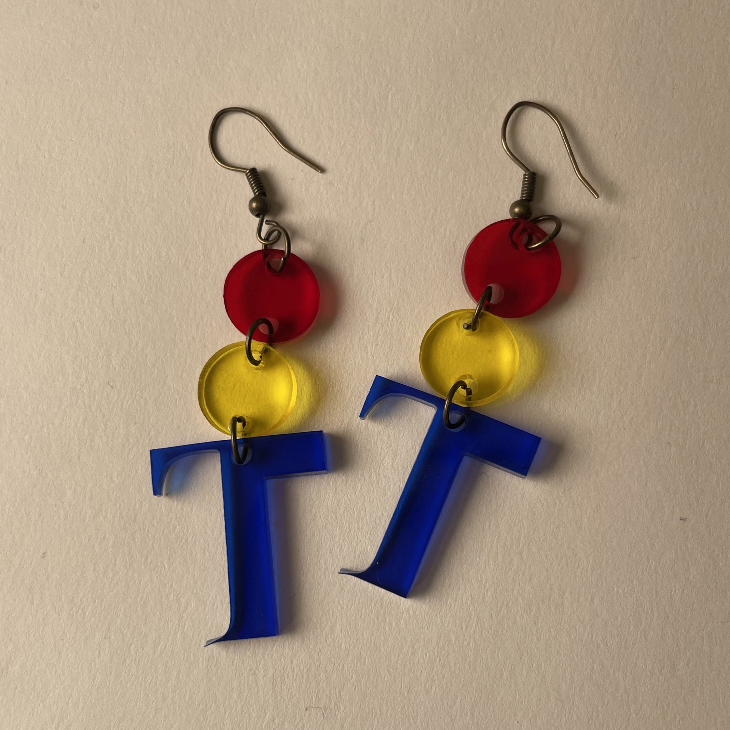 Two earrings that are laser-cut acrylic T shapes with two circle shapes added to the top of the T lay flat on a blank surface