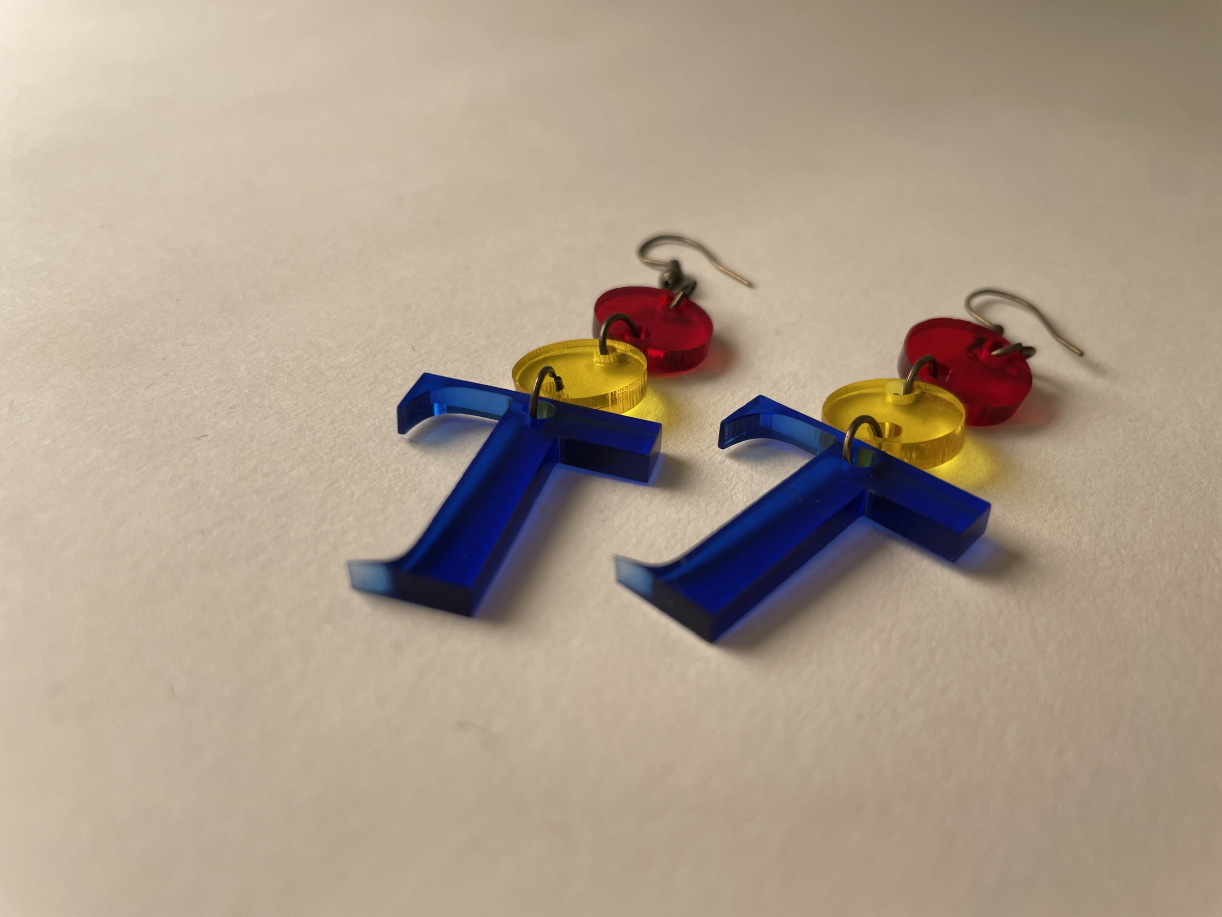 Two earrings that are laser-cut acrylic T shapes with two circle shapes added to the top of the T lay flat on a blank surface