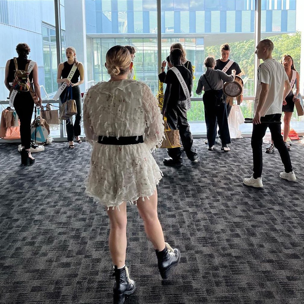 Models prepare backstage to line up for a fashion show, standing in front of a wall of floor-to-ceiling windows in the background