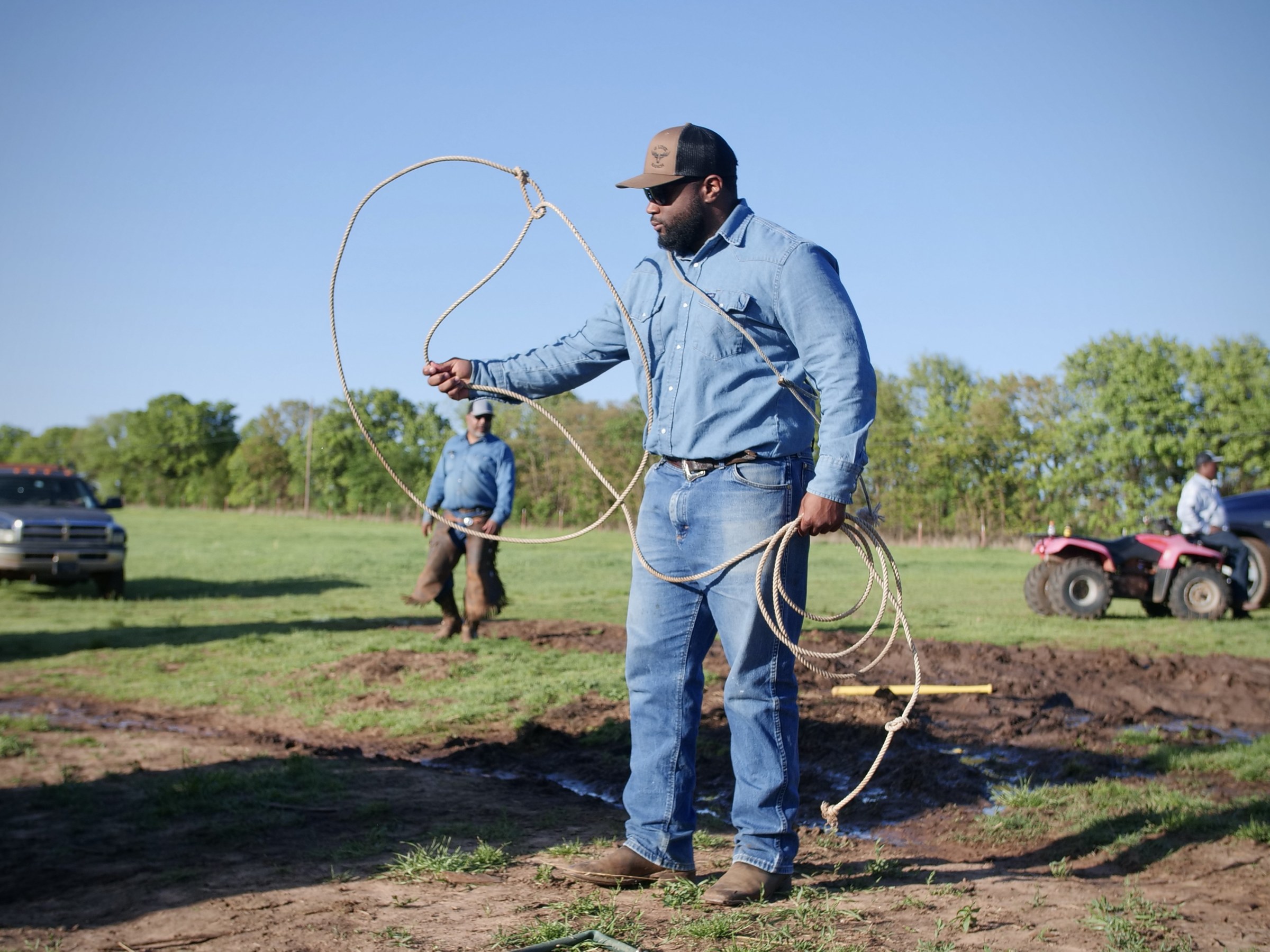 A man in denim pants and shirt twirls a lasso in the foreground, while a man rides a four-wheeler and another man walks in the background on a farm setting.