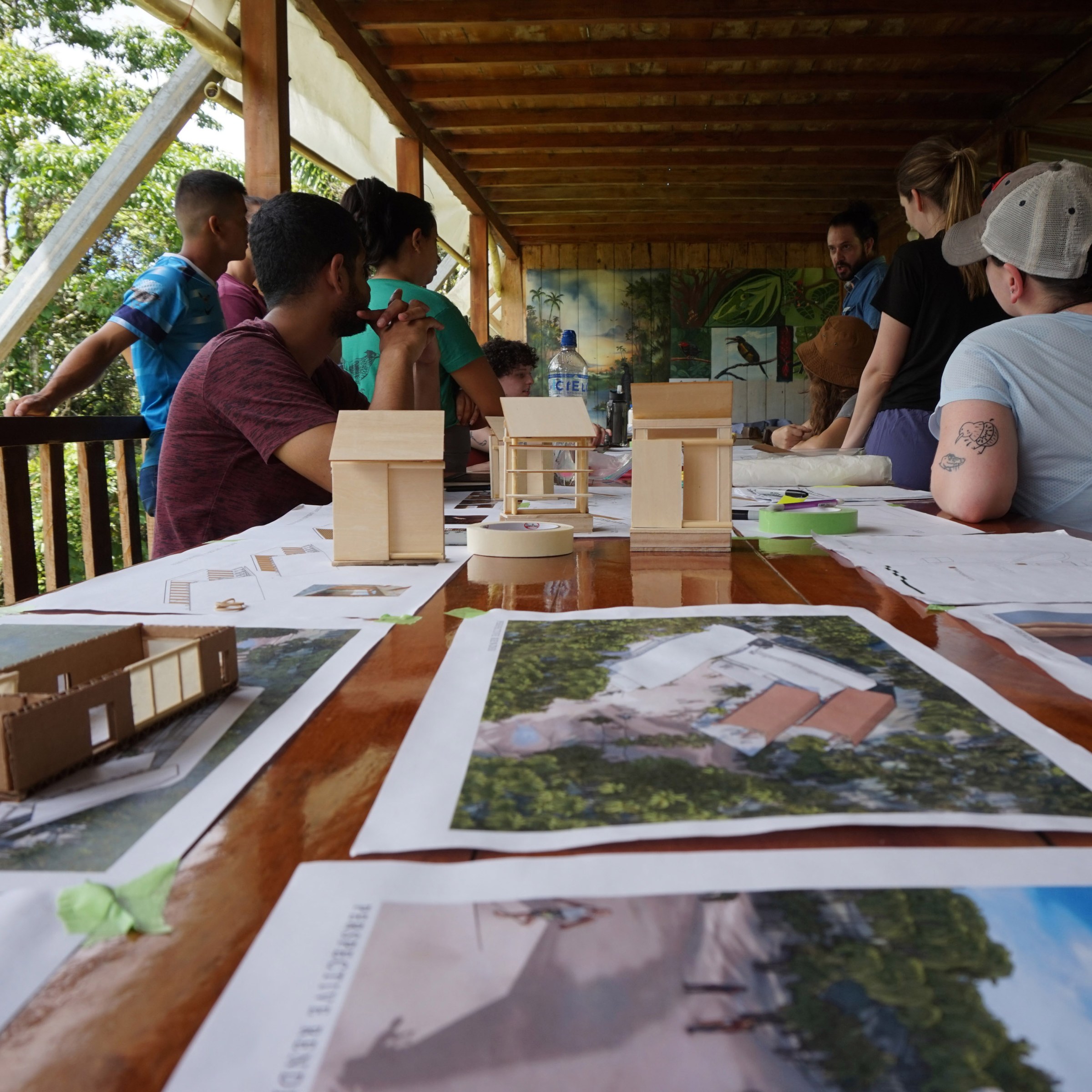 Architectural drawings and wood models cover a table in the foreground with students and people standing and sitting while talking in the background under a pavilion with a tropical forest on either side.