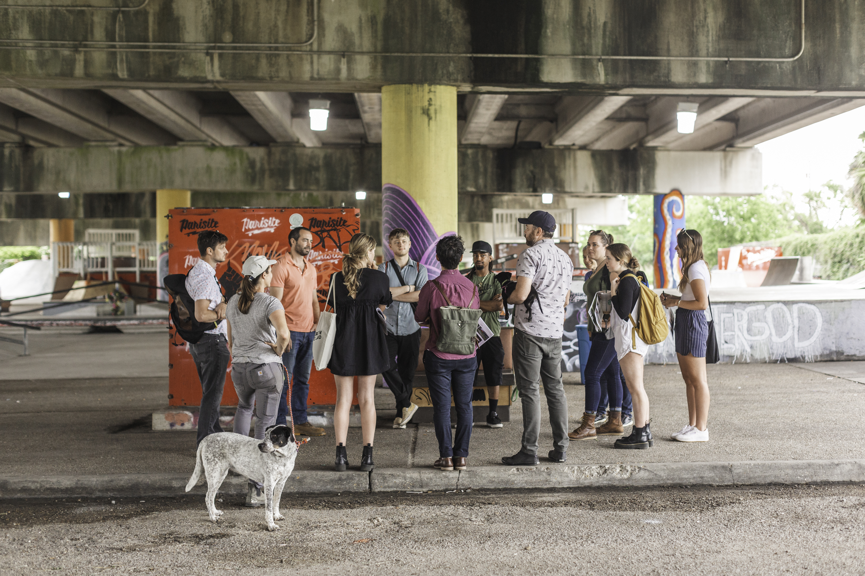 Photo of a group of students, faculty, and community partners standing and talking at Parisite, a concrete skatepark, under a highway overpass.