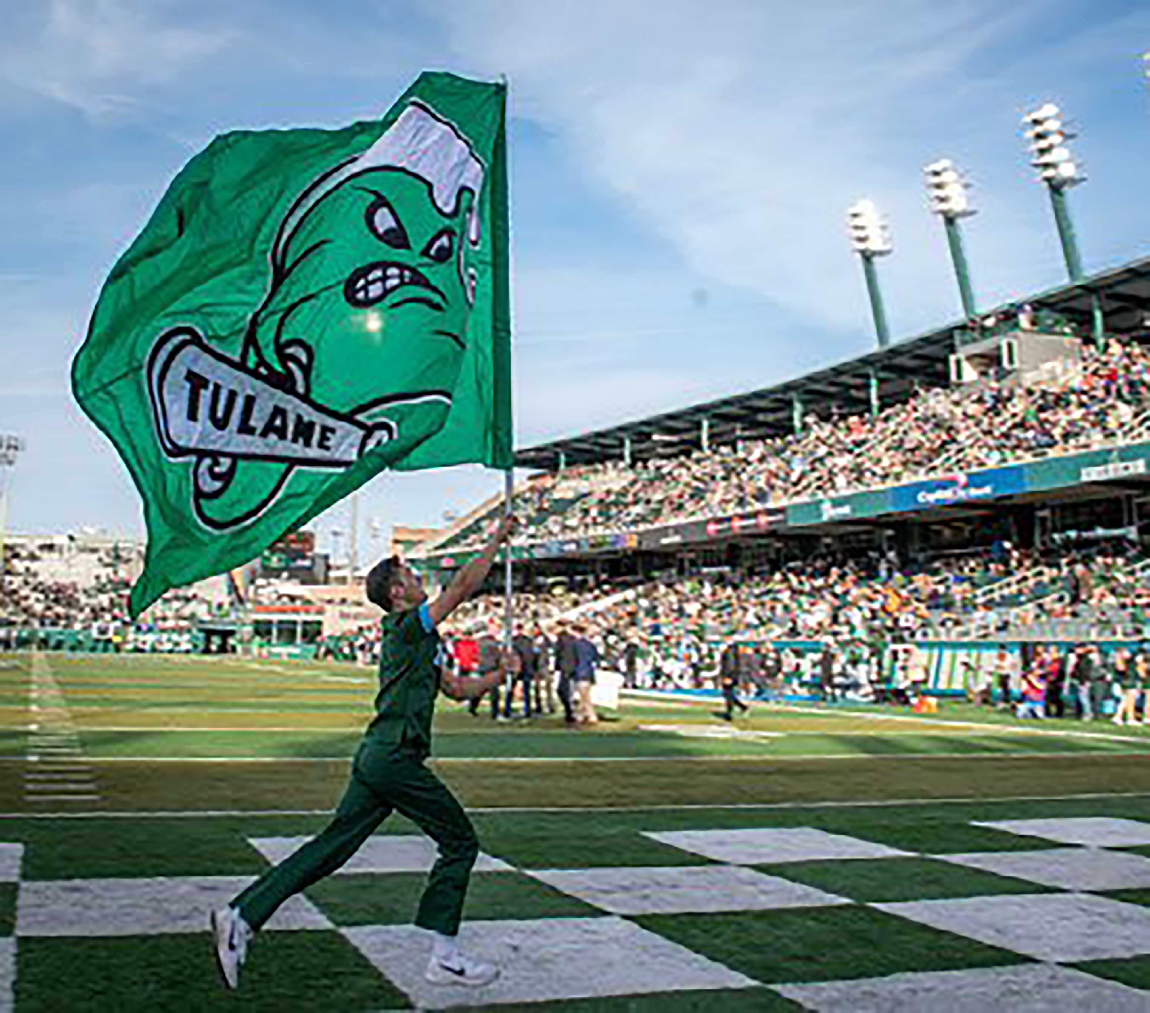 Photo of a man running across the endzone of a football stadium while holding up a large flag with Tulane's Angry Wave symbol on it; in the background are stadium stands full of people