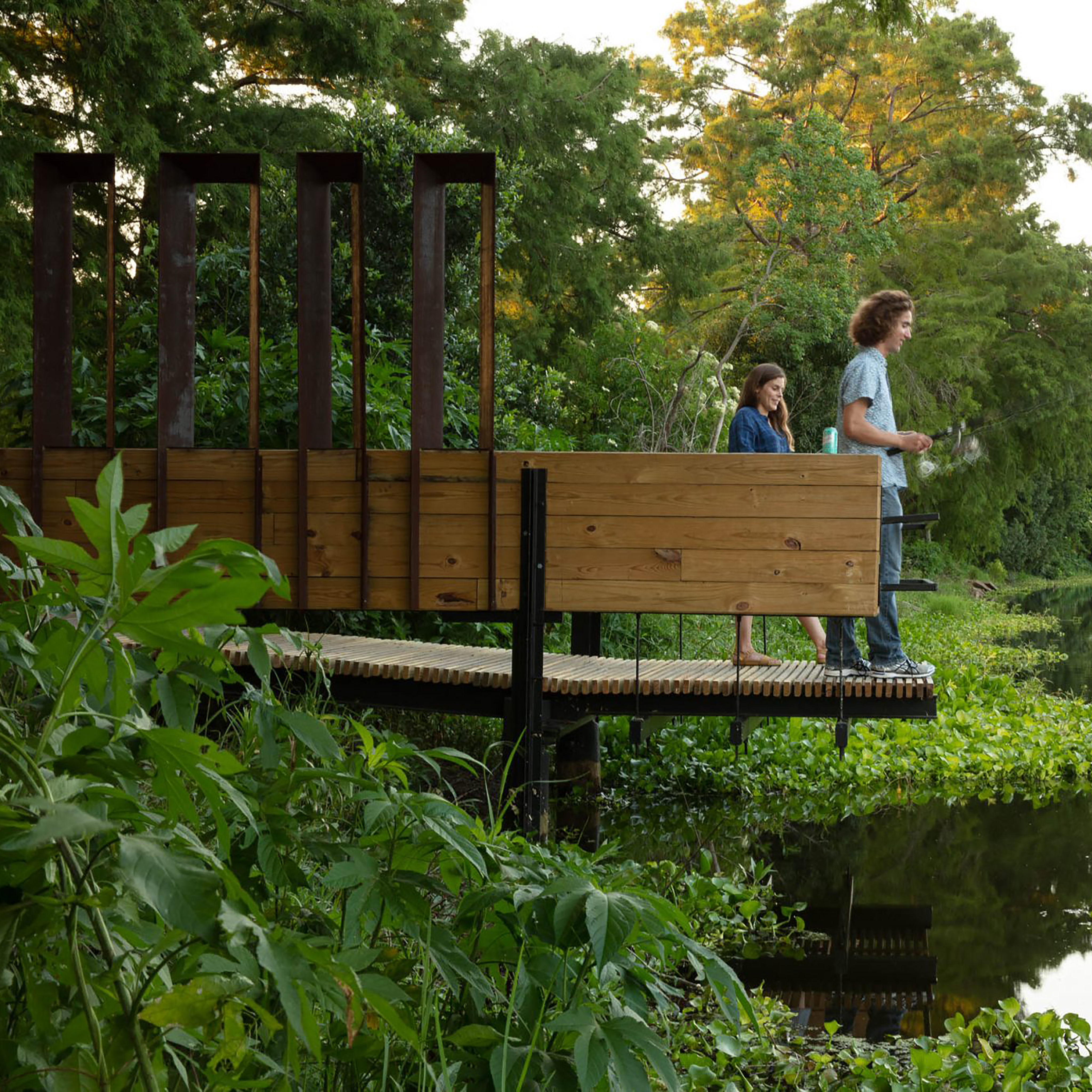 Photo of two people standing on a small pier made of steel and wooden beams and railing that is extended over a pond
