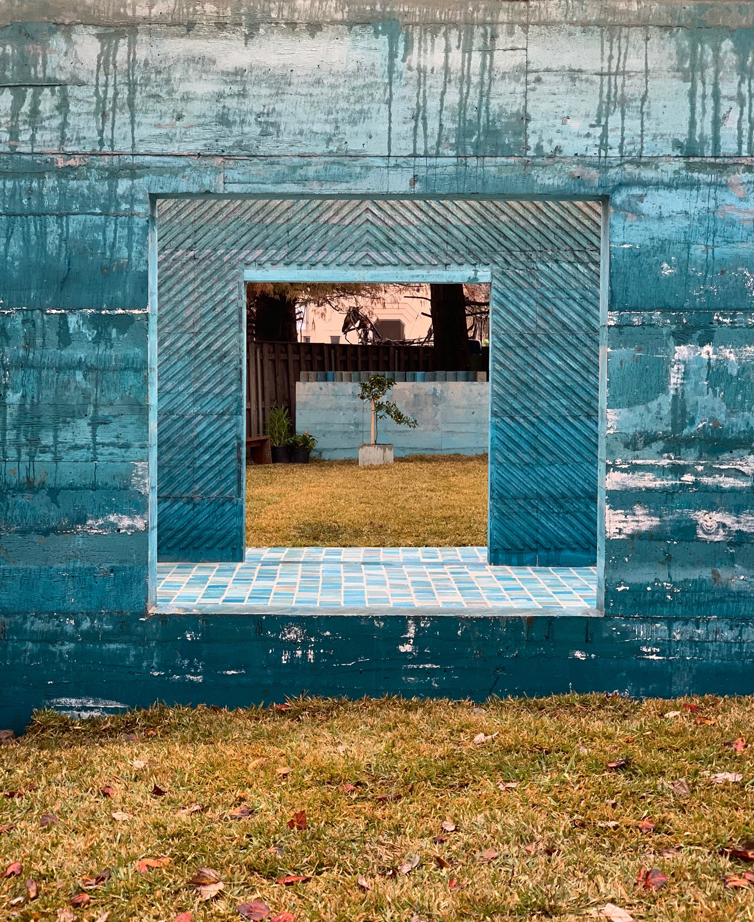 Photo outdoors looking directly through two square framed openings in concrete walls and a young tree in the background, framed by the two concrete window openings.