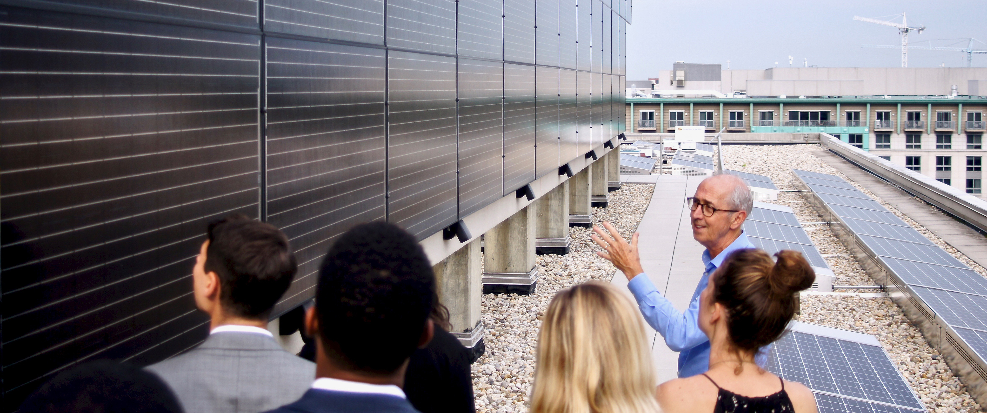 Photo of people standing on a rooftop and looking at a wall of solar panels to the left with solar panels on the rooftop to the right