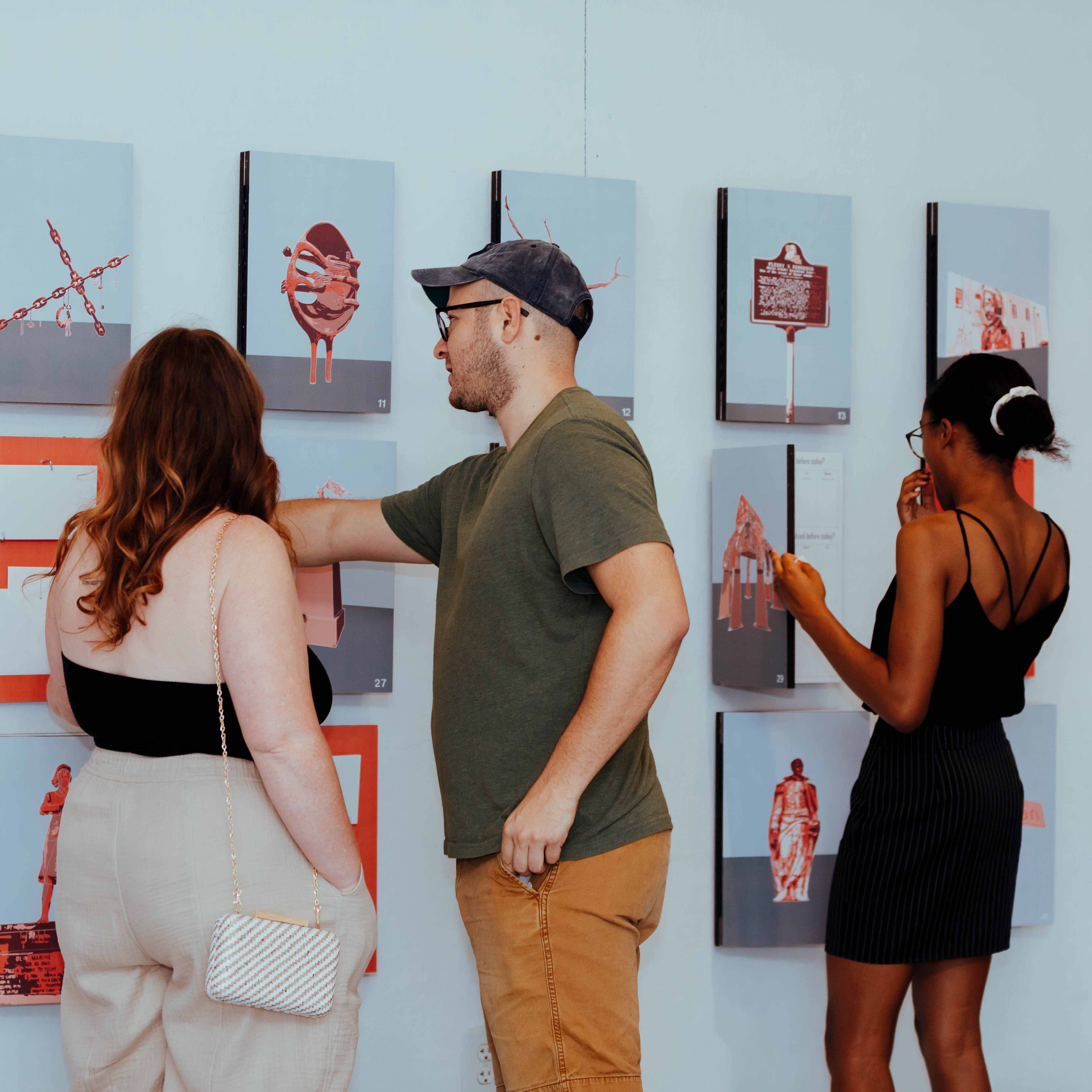 Three people standing with backs to the camera while looking at photos of monument features displayed on a wall at an exhibit
