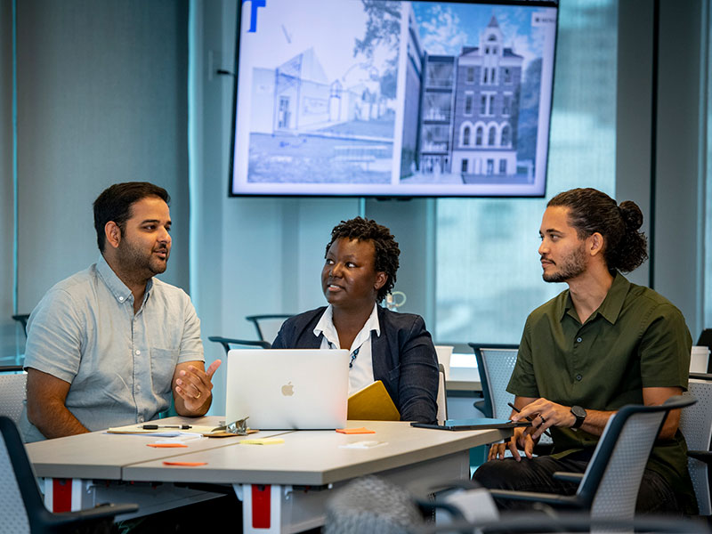 Three faculty sitting and talking at a table with a TV monitor screen in background showing architectural renderings