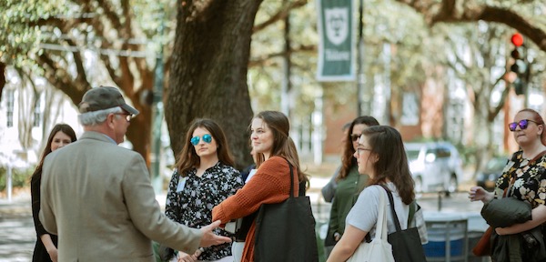 Man in beige blazer and green baseball cap speaking to a small group of students on the Tulane campus
