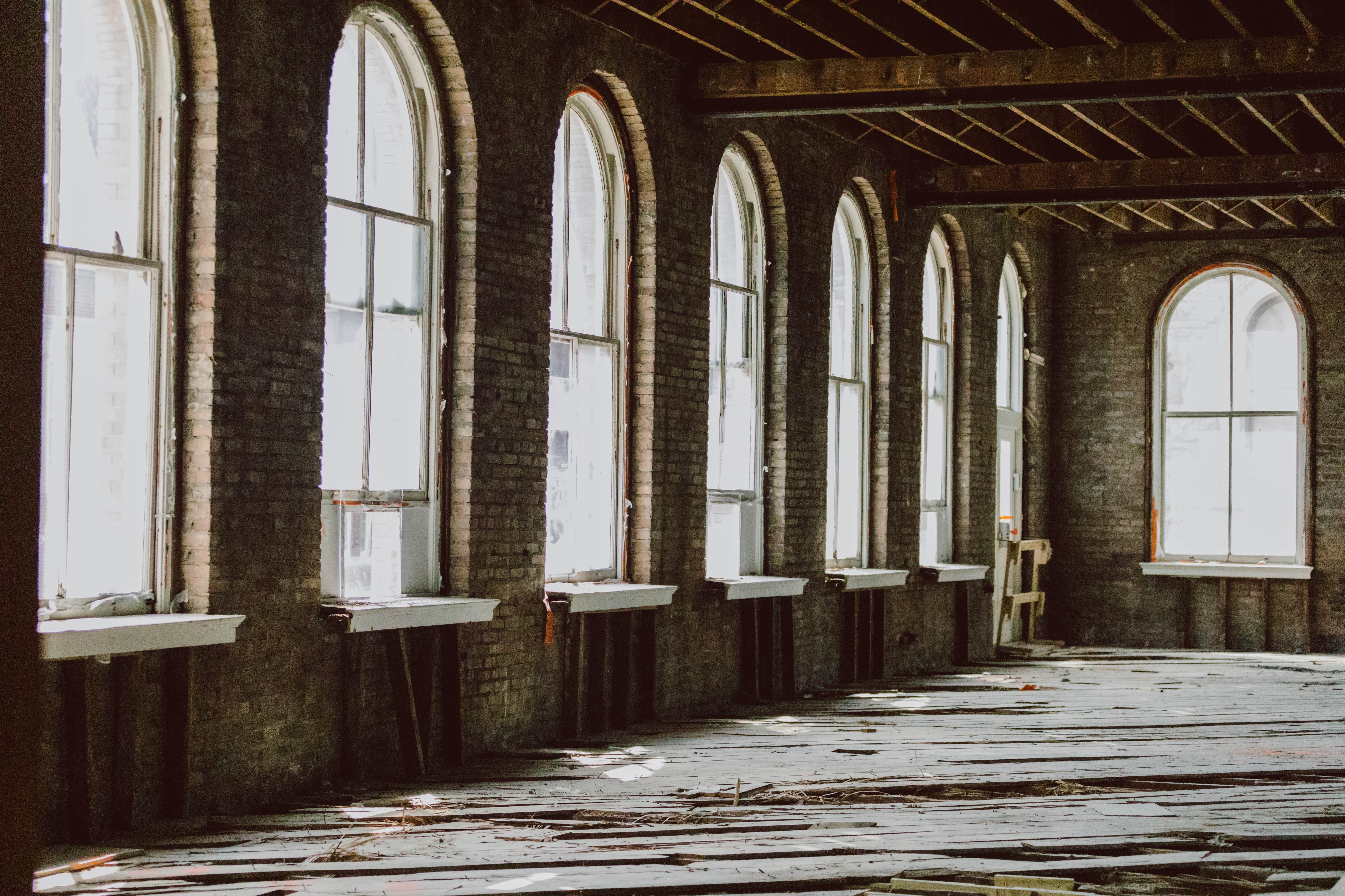 Interior of large room undergoing historic renovation with tall arched windows and exposed brick walls and wood ceiling and floors.