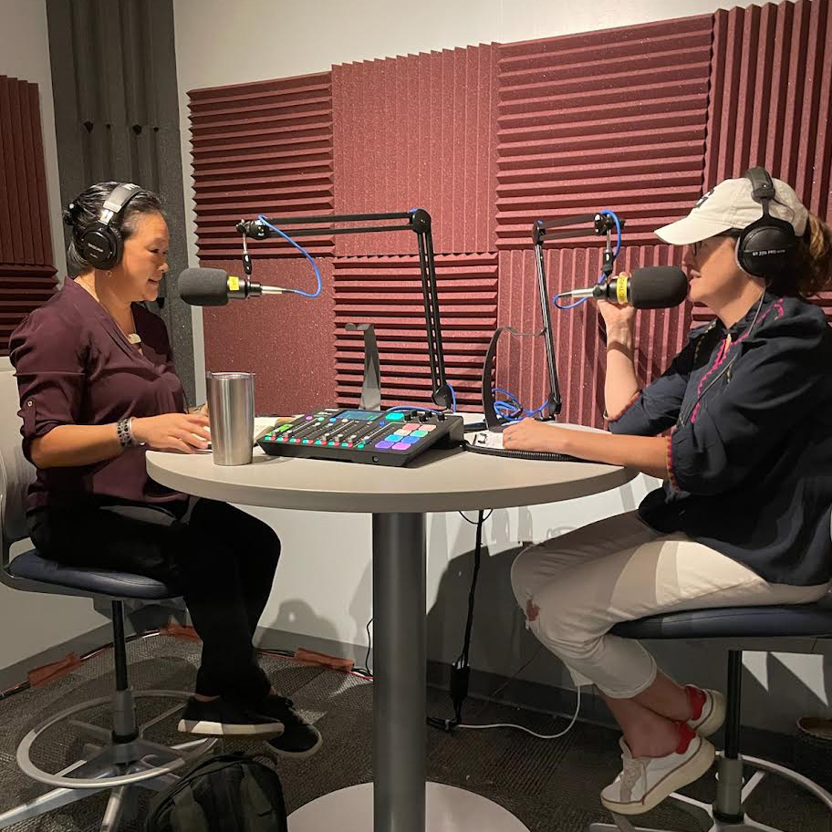 Two women, wearing headphones and speaking into large microphones, are sitting on either side of a round table inside a sound booth