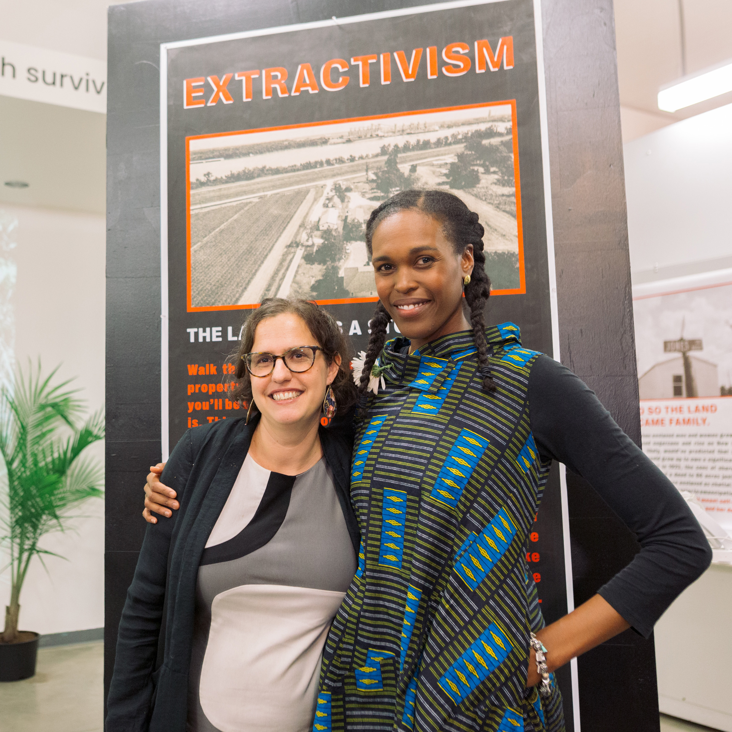 Two women smiling with arms around each others shoulders pose in front of an exhibition poster with the title "Extractivism" written on the top