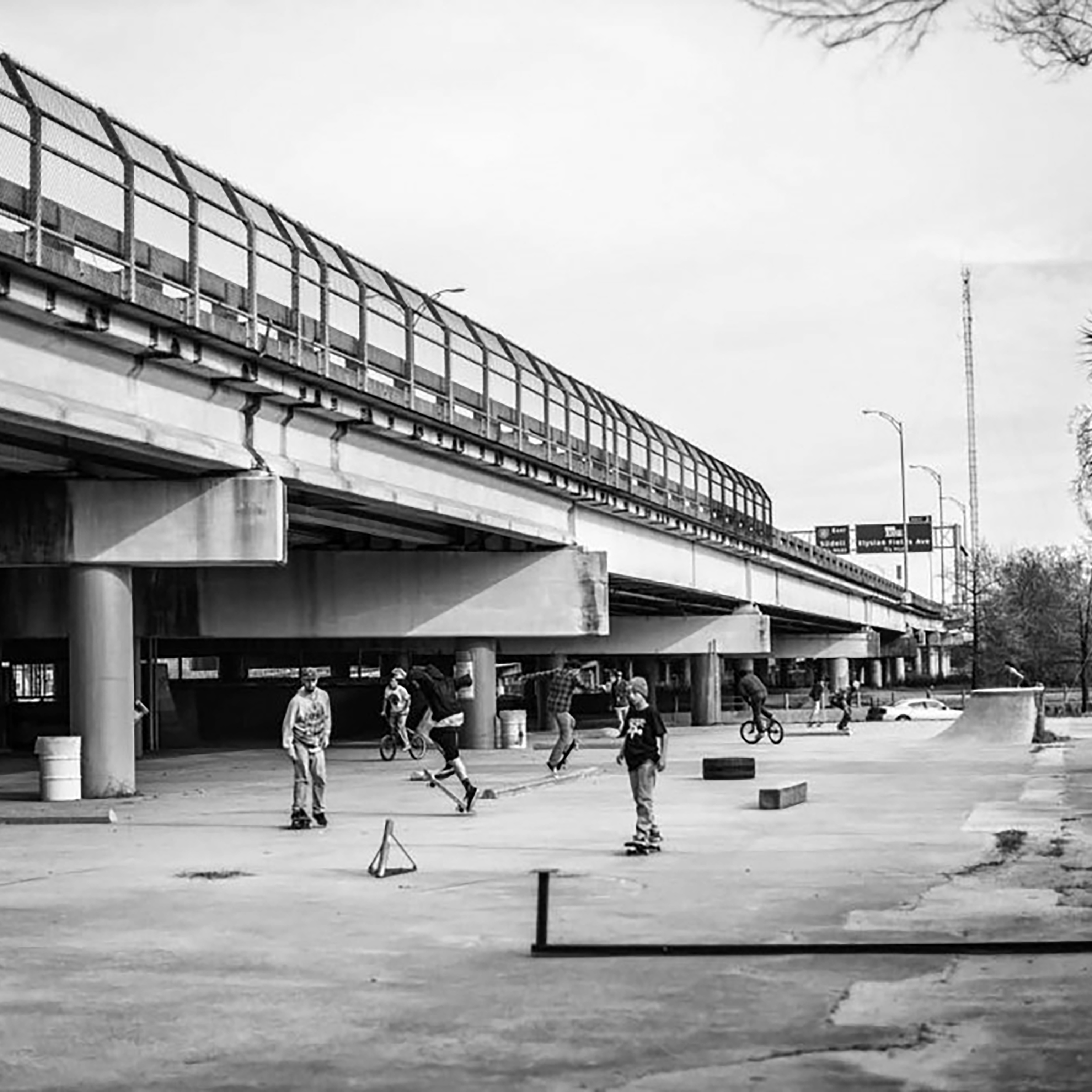Black and white photo of group of people skateboarding under an overpass