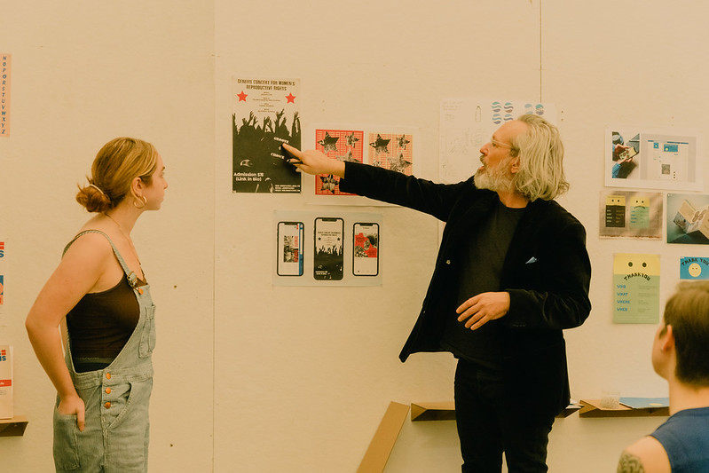 Image of a design class final review, with the student's work pinned up to the wall, while the student and the teacher discuss the work on the board.