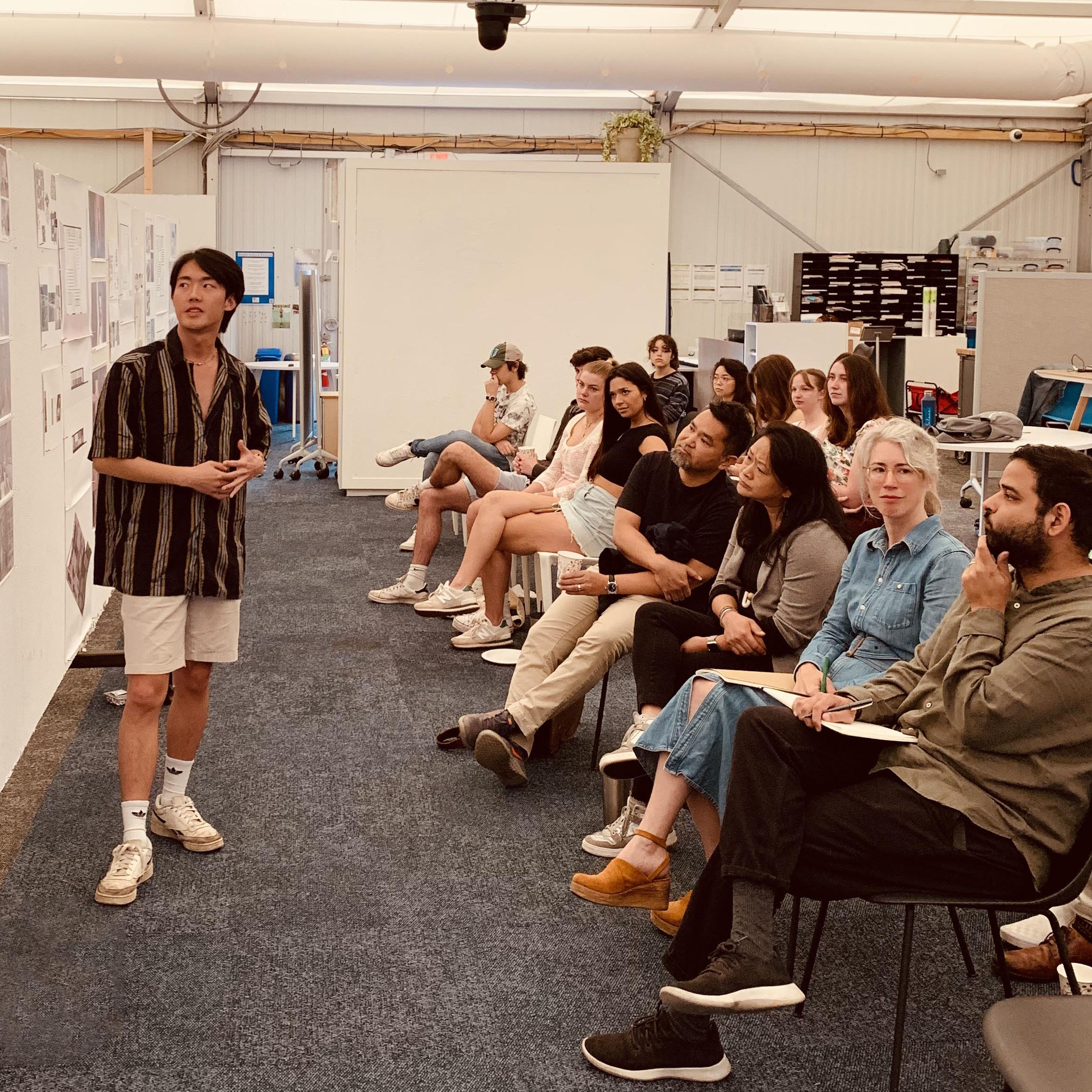 Student stands next to and looks at pinned-up prints on the left side of the frame, while a long row of reviewers seated in chairs look at the work on the right side of the frame. 