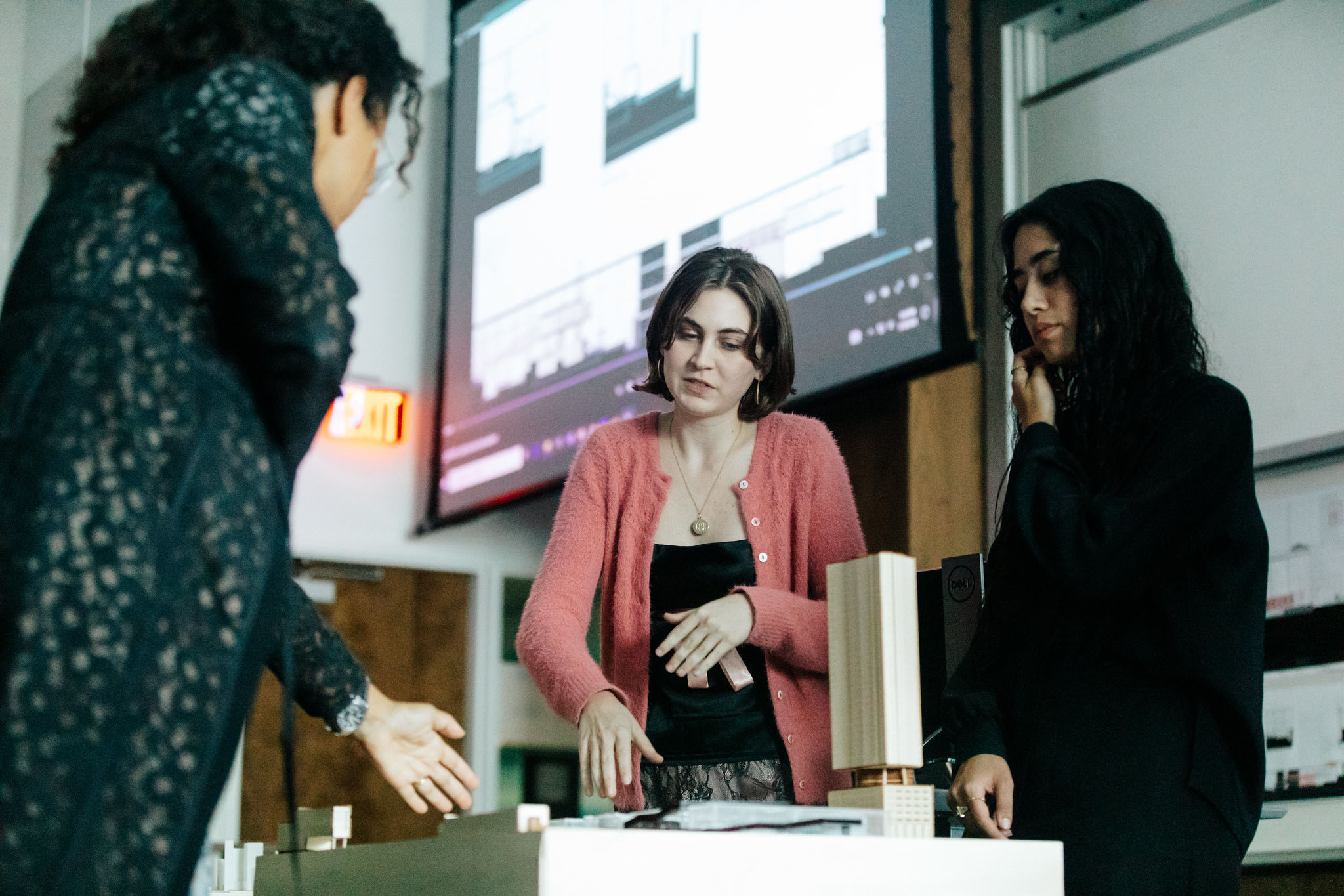Two students engage in discussion with a professor looking down at a model on a table with a projected presented on a screen in the background.