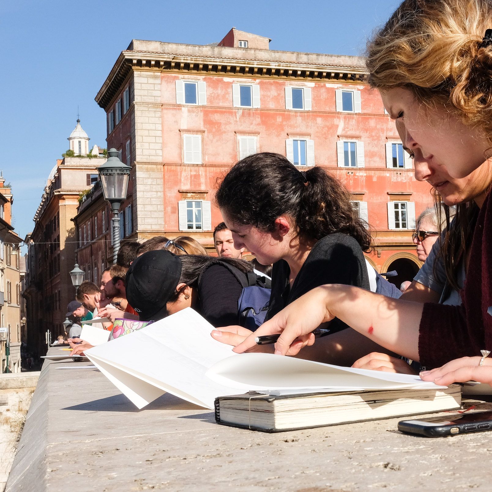 close up, looking up at a row of students doing hand drawings with sketchbooks on top of a stone wall with a historic 4-story building in the background.