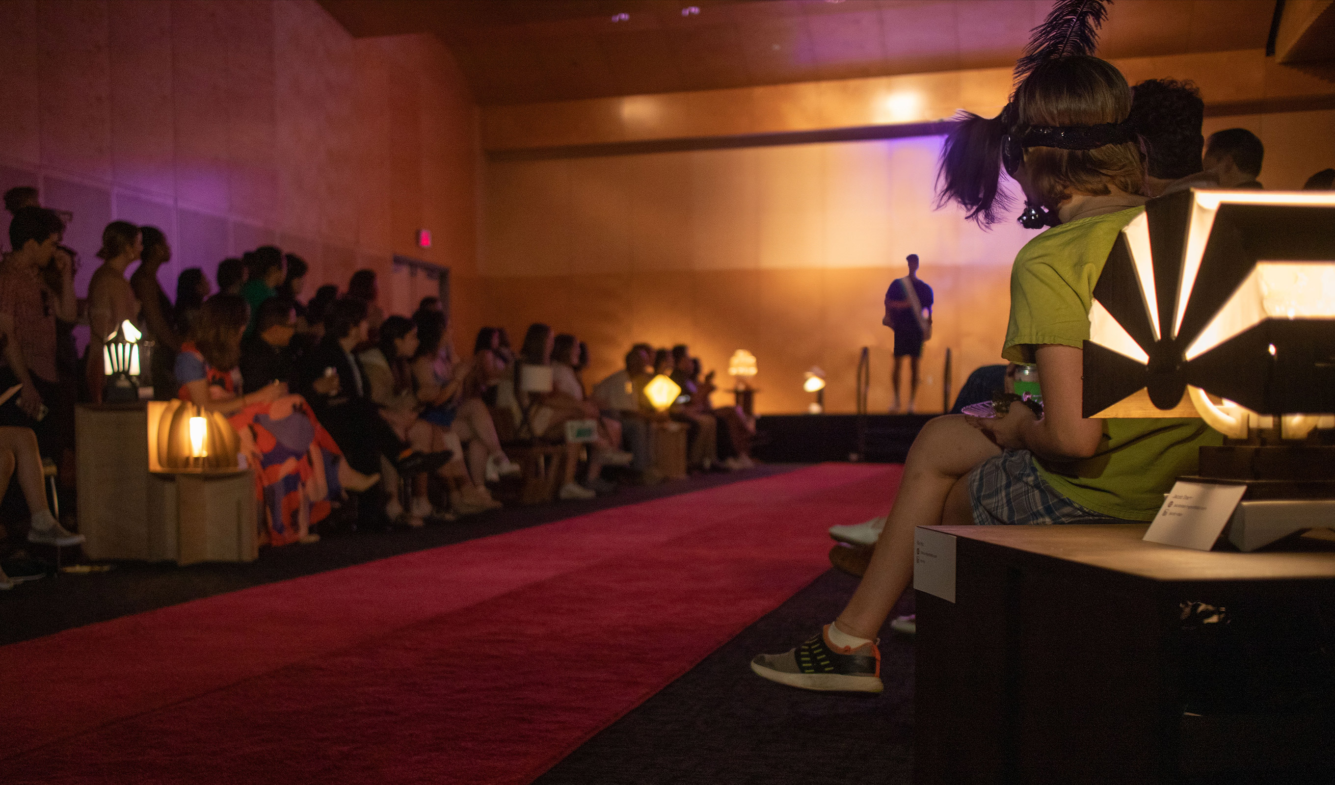 View of a red carpet runway inside a darkened room with dozens of people seated and standing on either side of the runway while a model stands in the background, getting ready to walk down the runway.