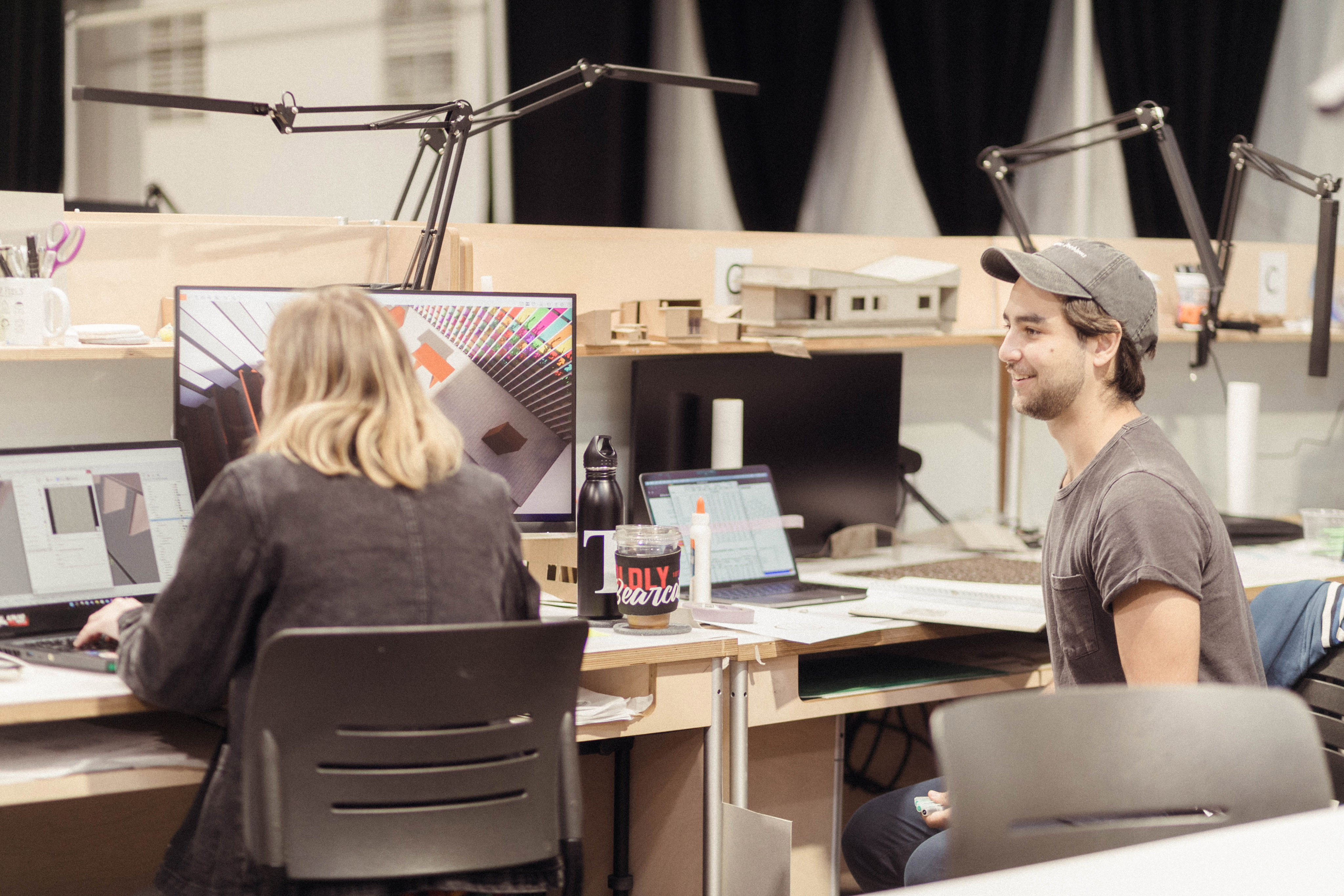 View from behind the backs of two students, one smiling, sitting side by side in front of wooden studio desks with computers and monitors.