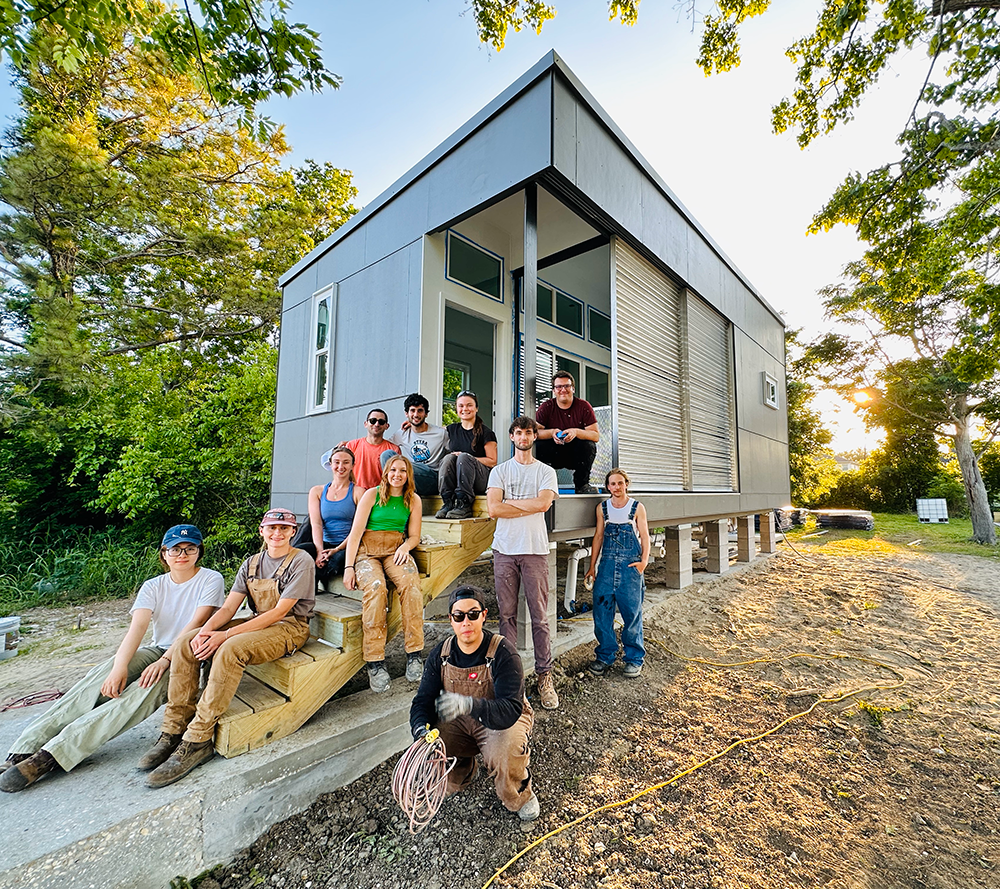 Group photo of students sitting and standing in front of a tiny house built for a homeless man lots of trees around the edges of the home.