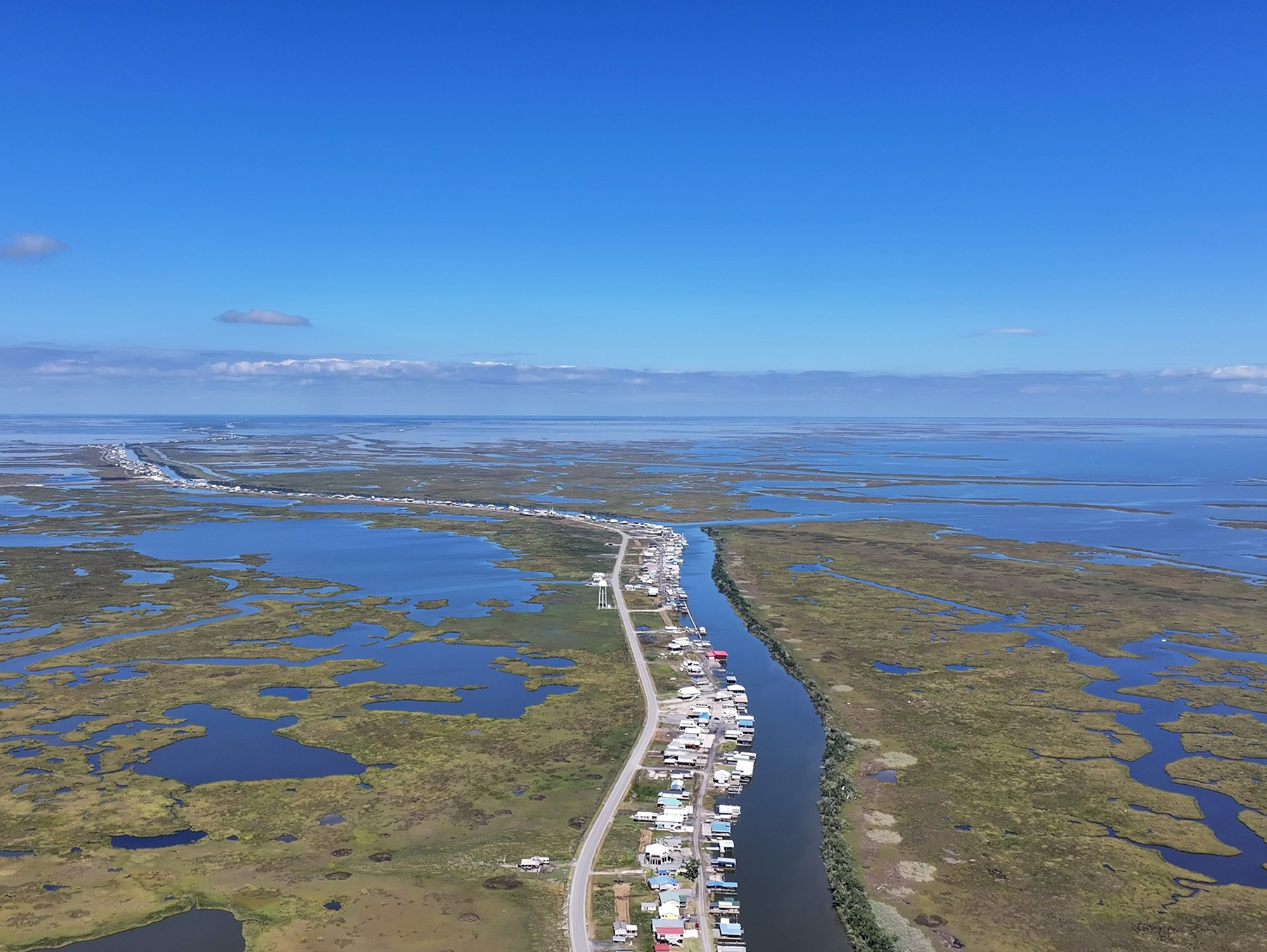 Aerial photo of a winding bayou, the left side lined with a single street of homes surrounded by sparse wetlands stretched out into the horizon.