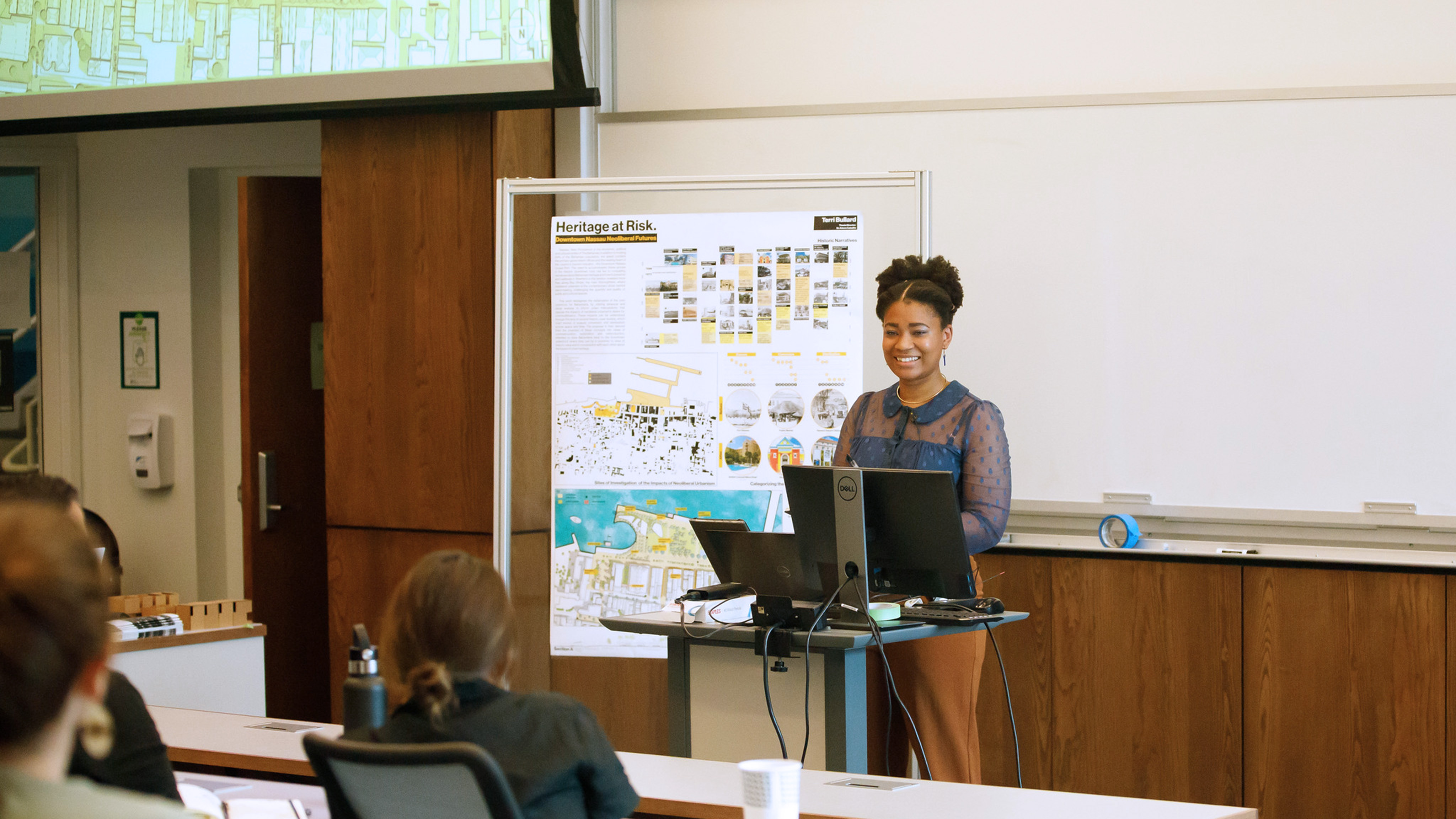 A student smiles while presenting their work at the front of a classroom, standing behind a podium.