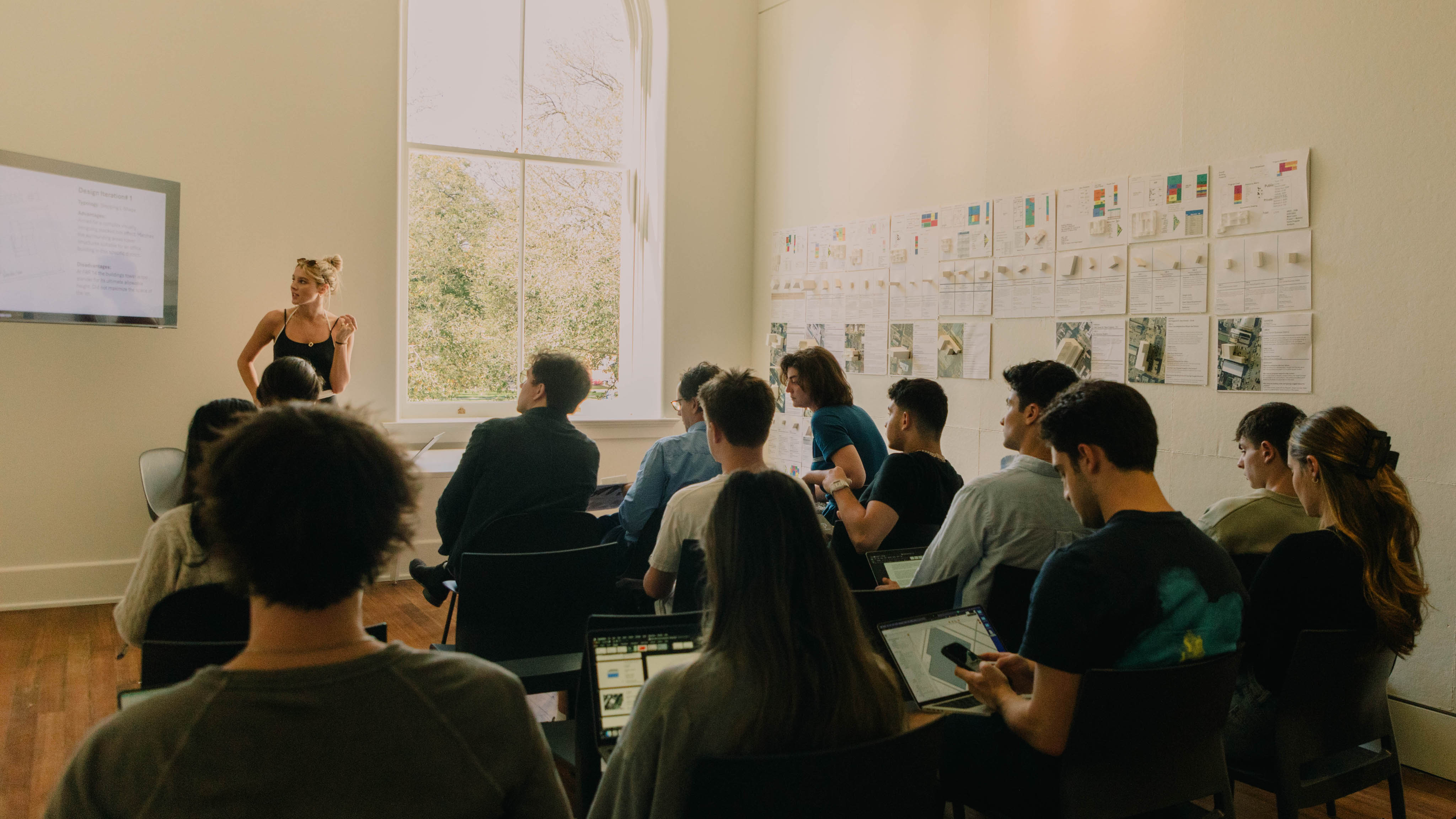 A group sits in chairs with back to camera while a student in the background stands next to a tall window and presents on a digital screen
