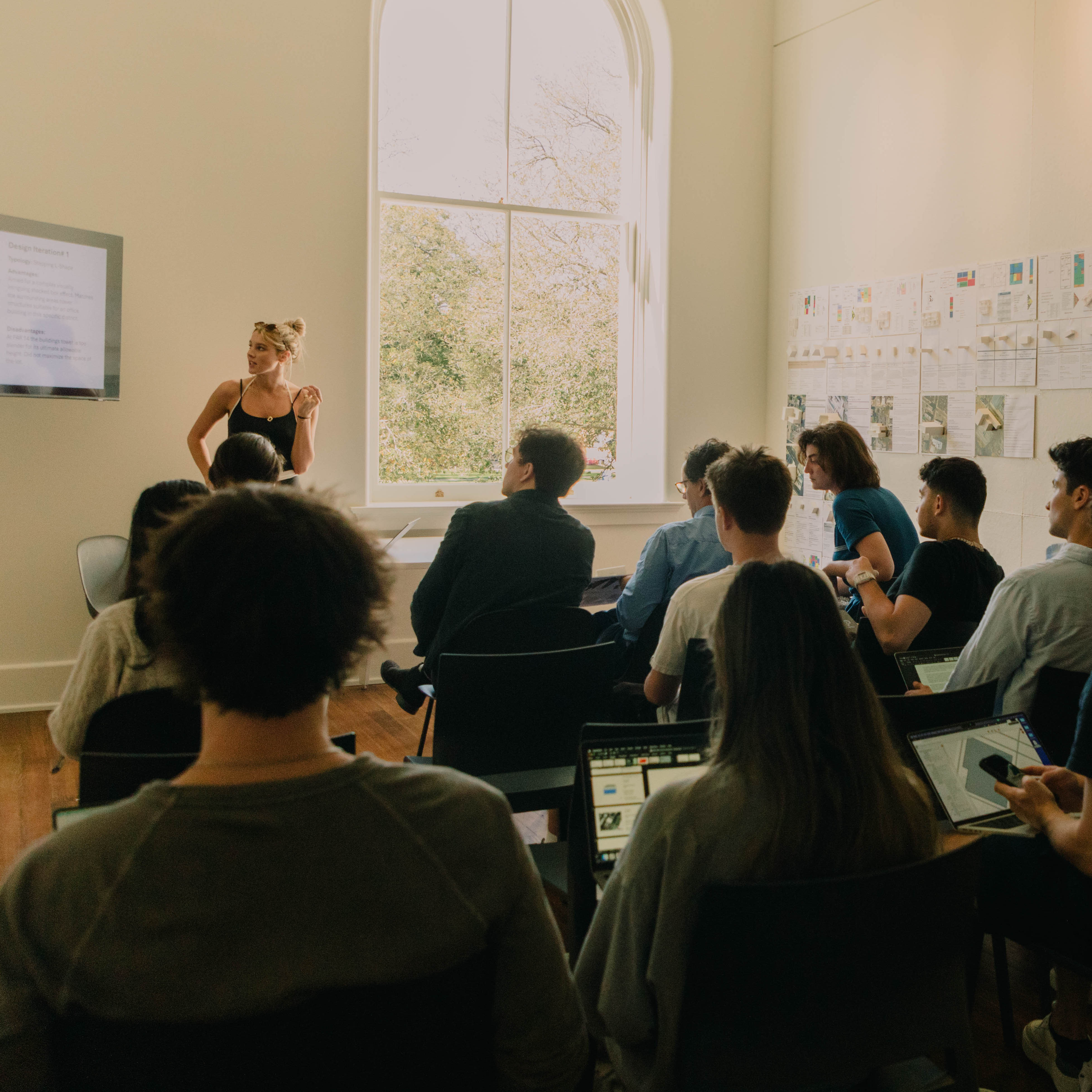 Student presents their work standing next to a tall open window in background, while rows of reviewers and other students with backs to camera in foreground listen.