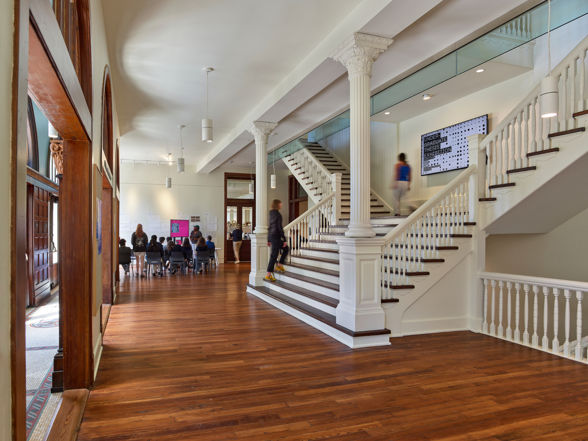 Inside corner view of historic Favrot Lobby with a large, Y-shaped staircase in the middle and students presenting work in the background.