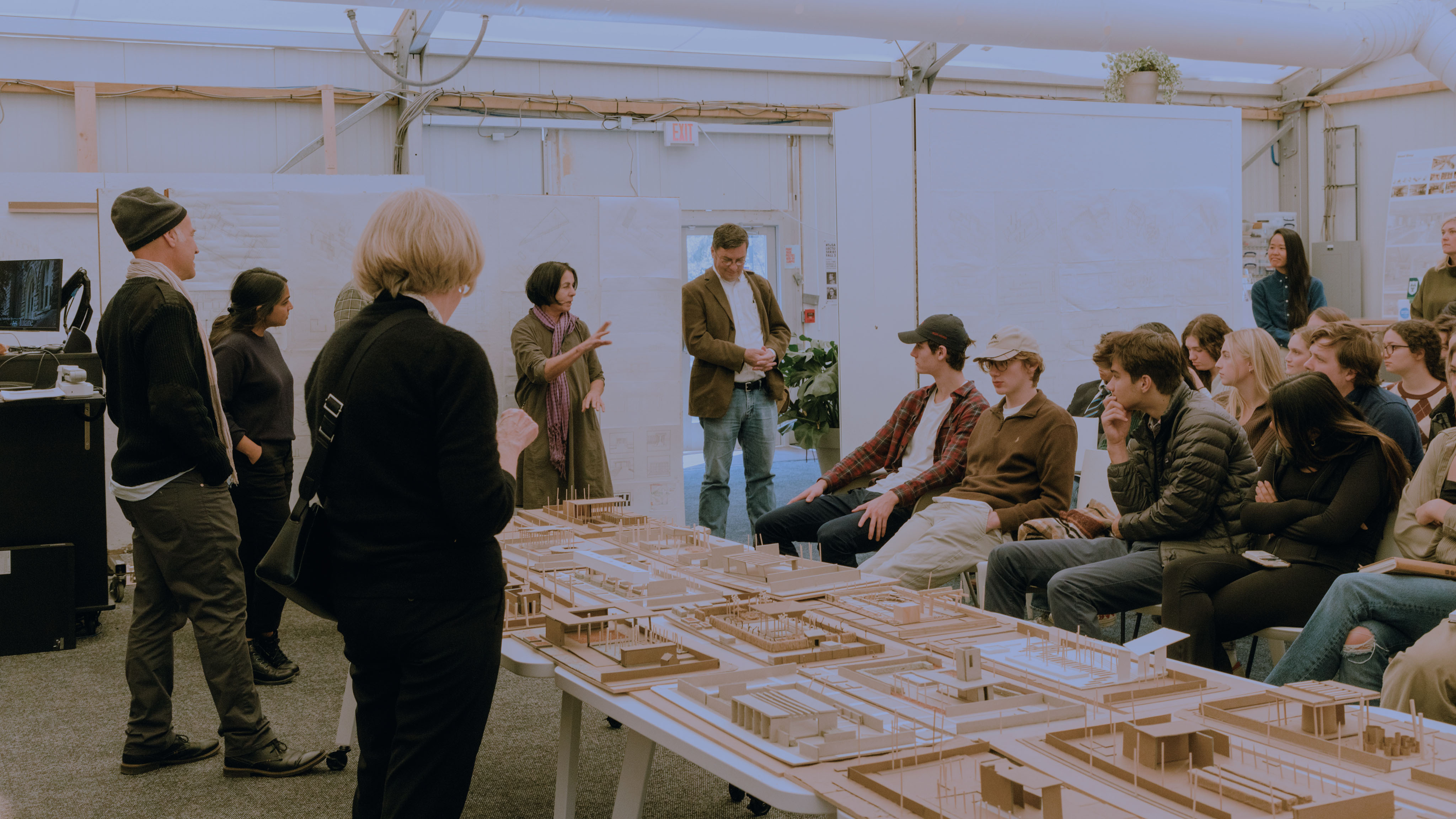 Students sit to the right and faculty stand to the left, looking at a large site model on top of a table in the middle of an open room.