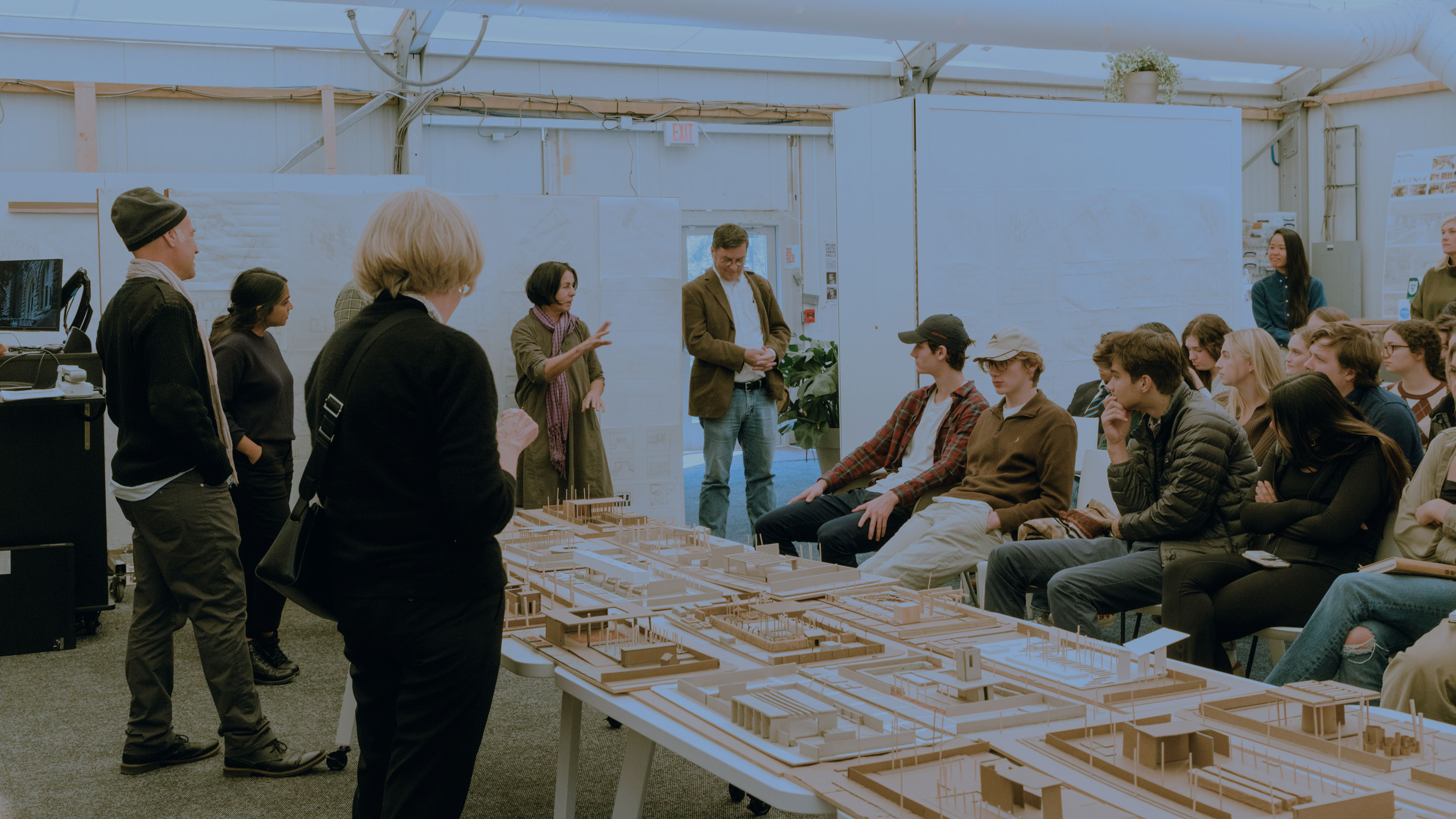 Students sit to the right and faculty stand to the left, looking at a large site model on top of a table in the middle of an open room.