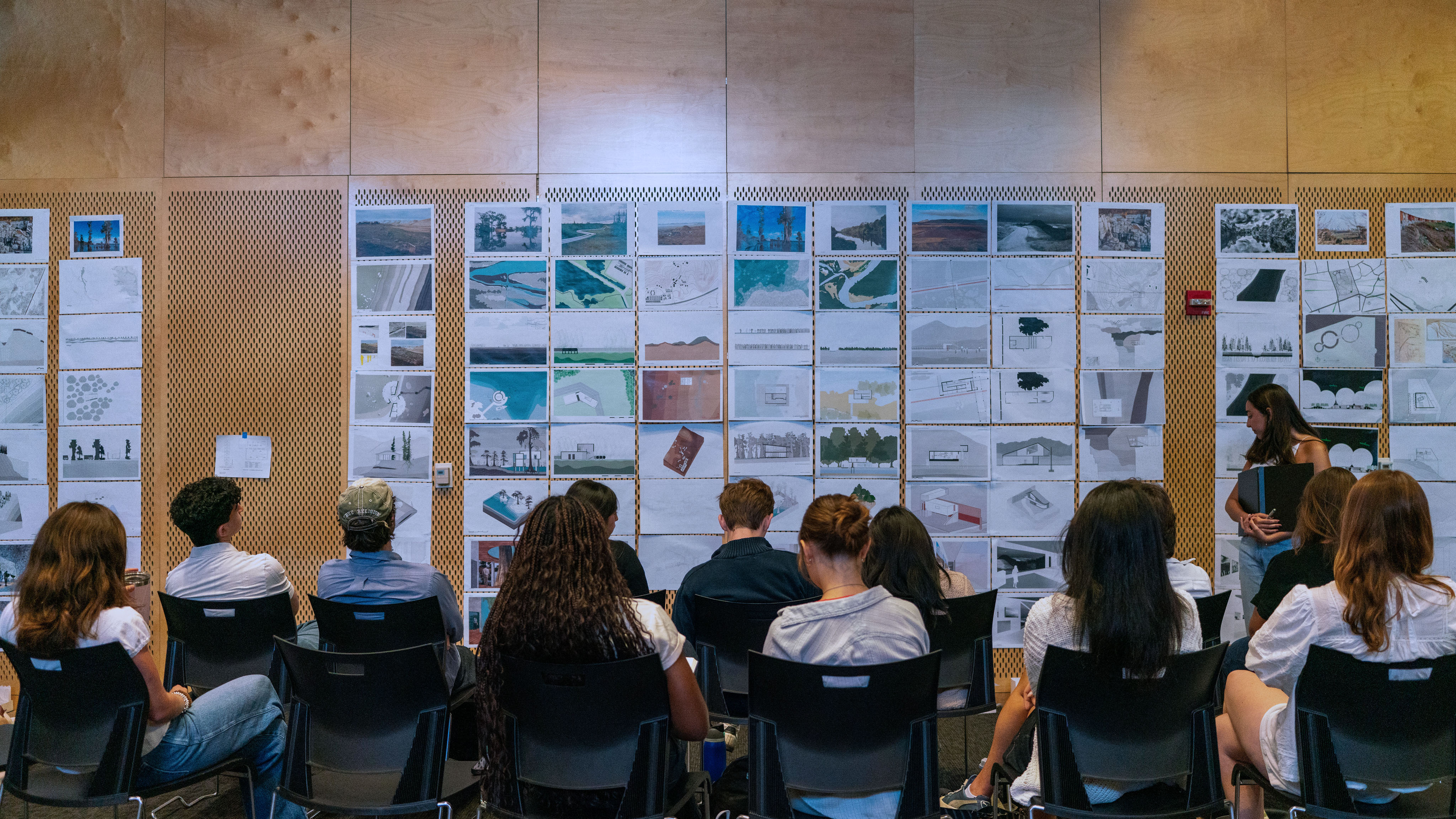 View from behind the heads of students sitting, facing a wall of pinned up drawings and designs.
