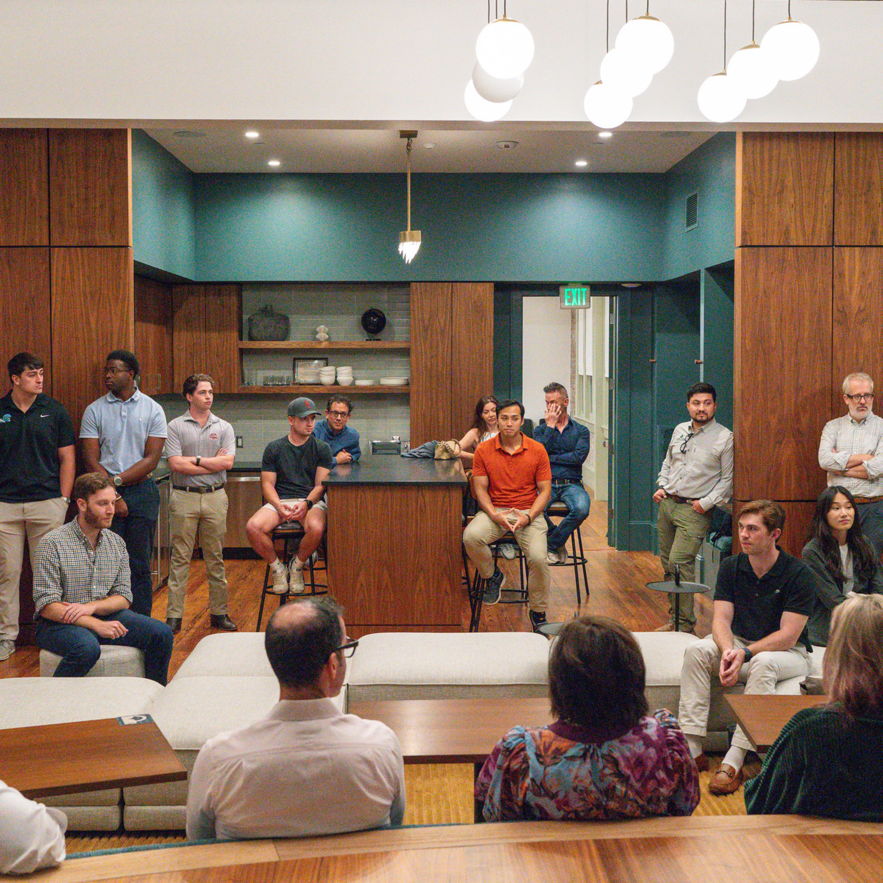 Students and faculty sit and stand while listening to someone talk on the right side in an open layout kitchen-dining area of a real estate development project.
