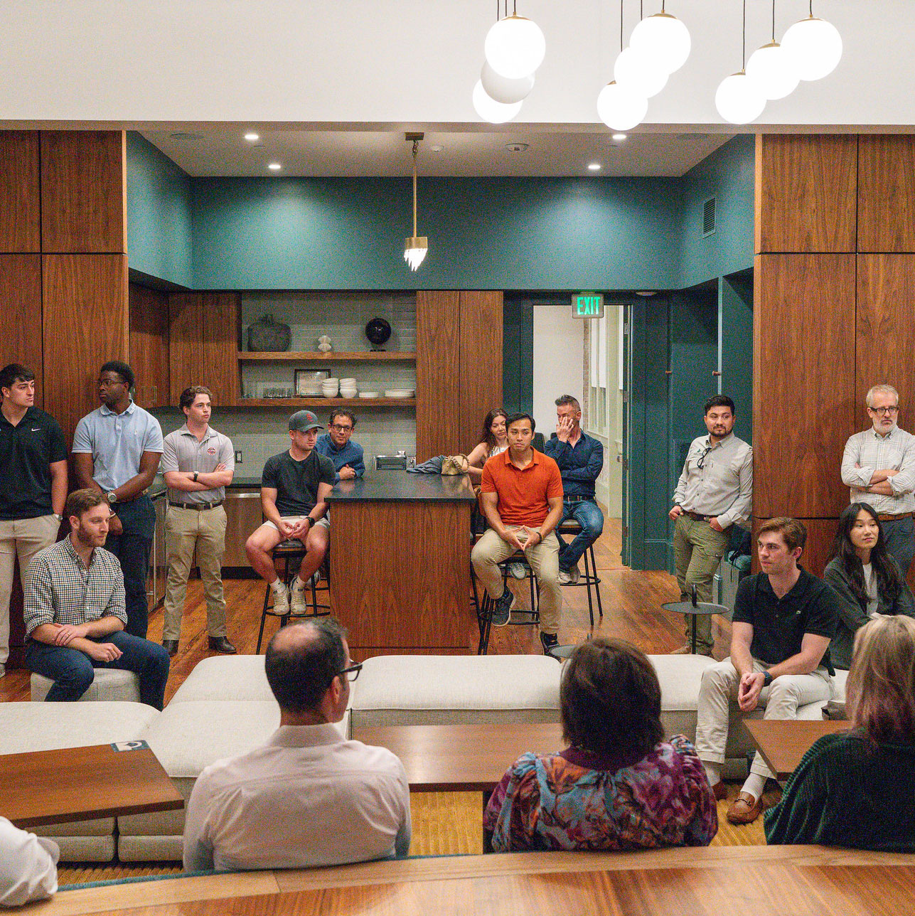 Students and faculty sit and stand while listening to a faculty talk on the right side in an open layout kitchen-dining area of a real estate development project.