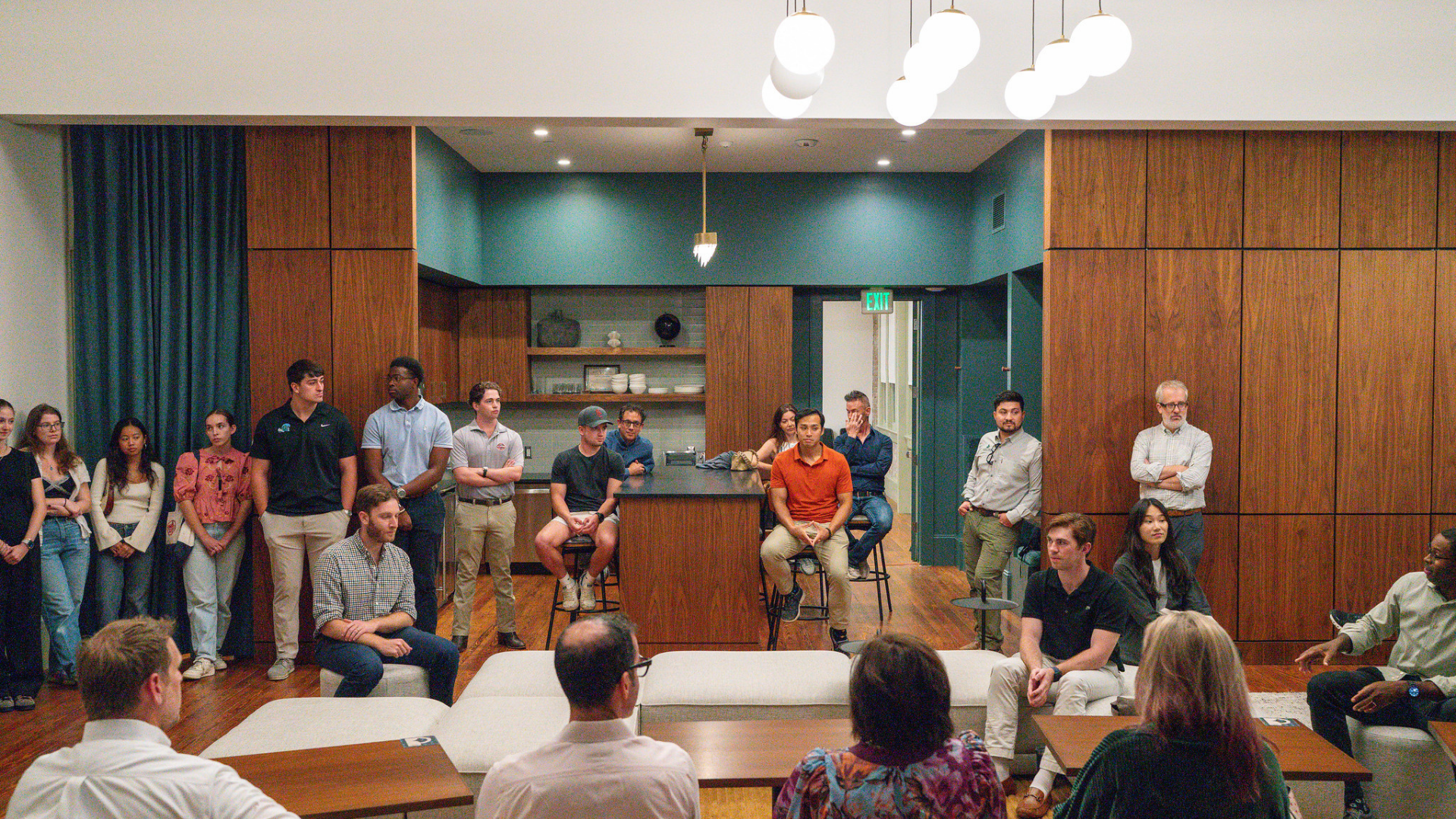 Students and faculty sit and stand while listening to a faculty talk on the right side in an open layout kitchen-dining area of a real estate development project.