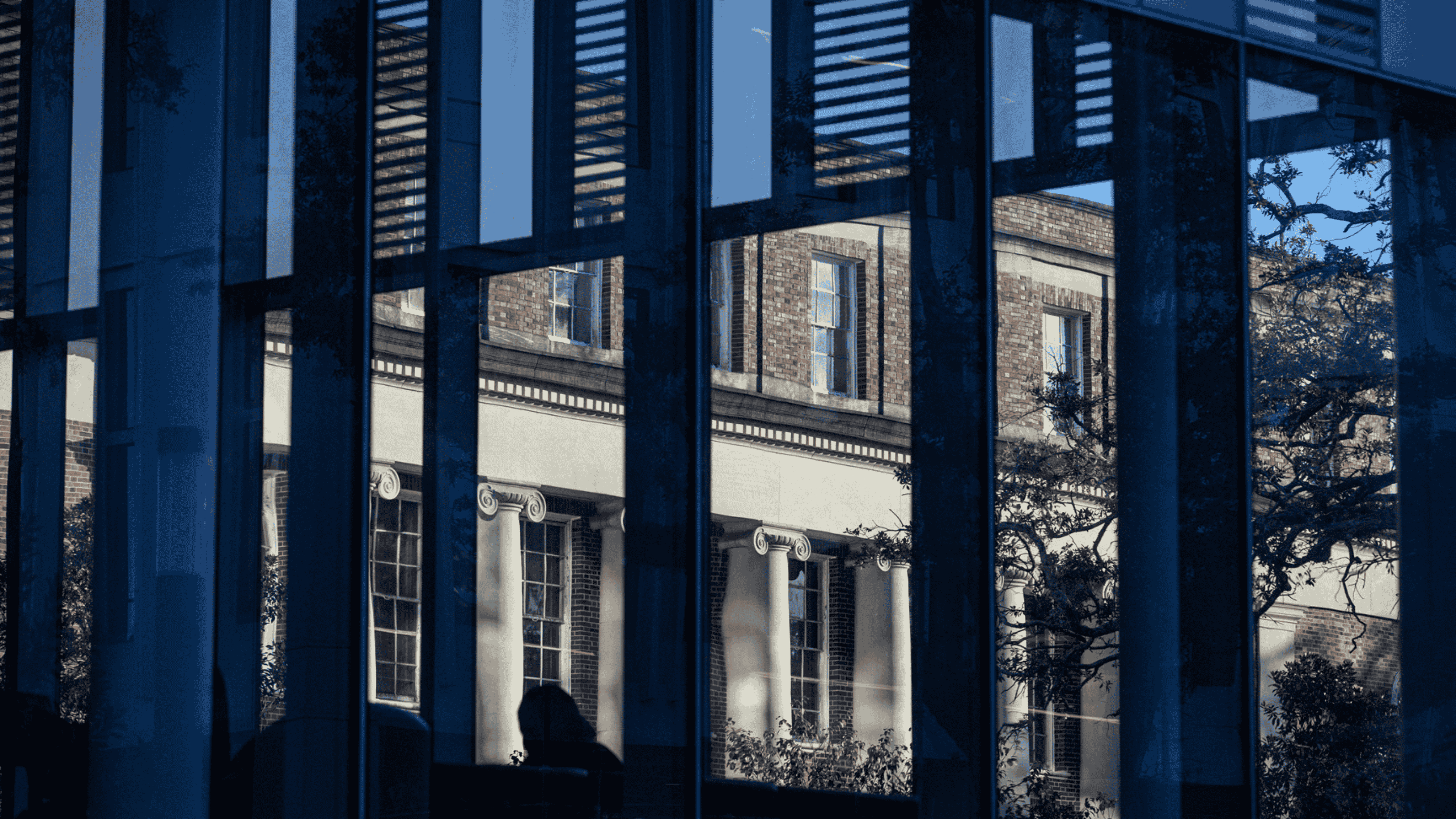 An historic stone building is shown in the angled reflection of a mirror-windowed modern building.