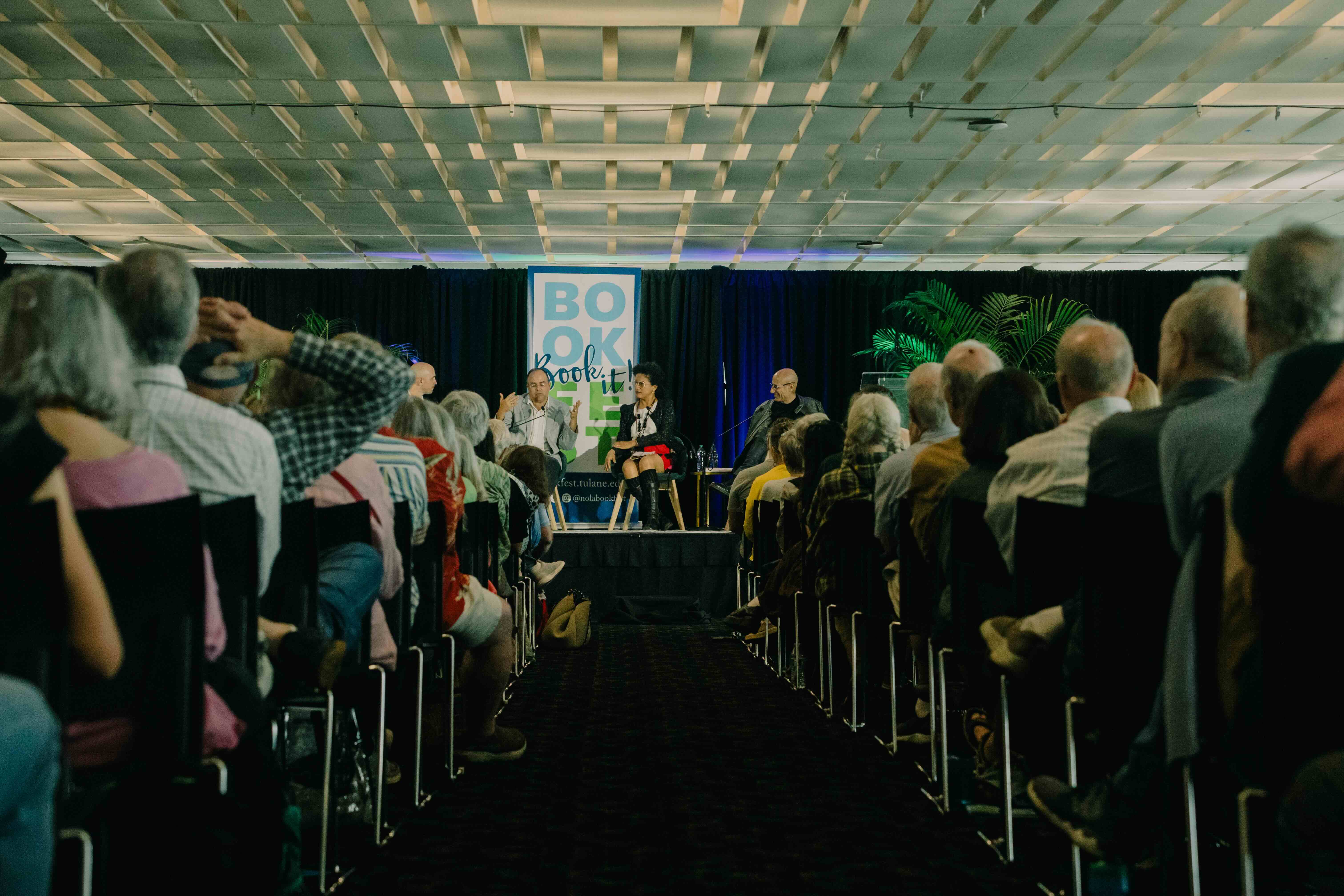 four people sit on a dais speaking surrounded by an audience