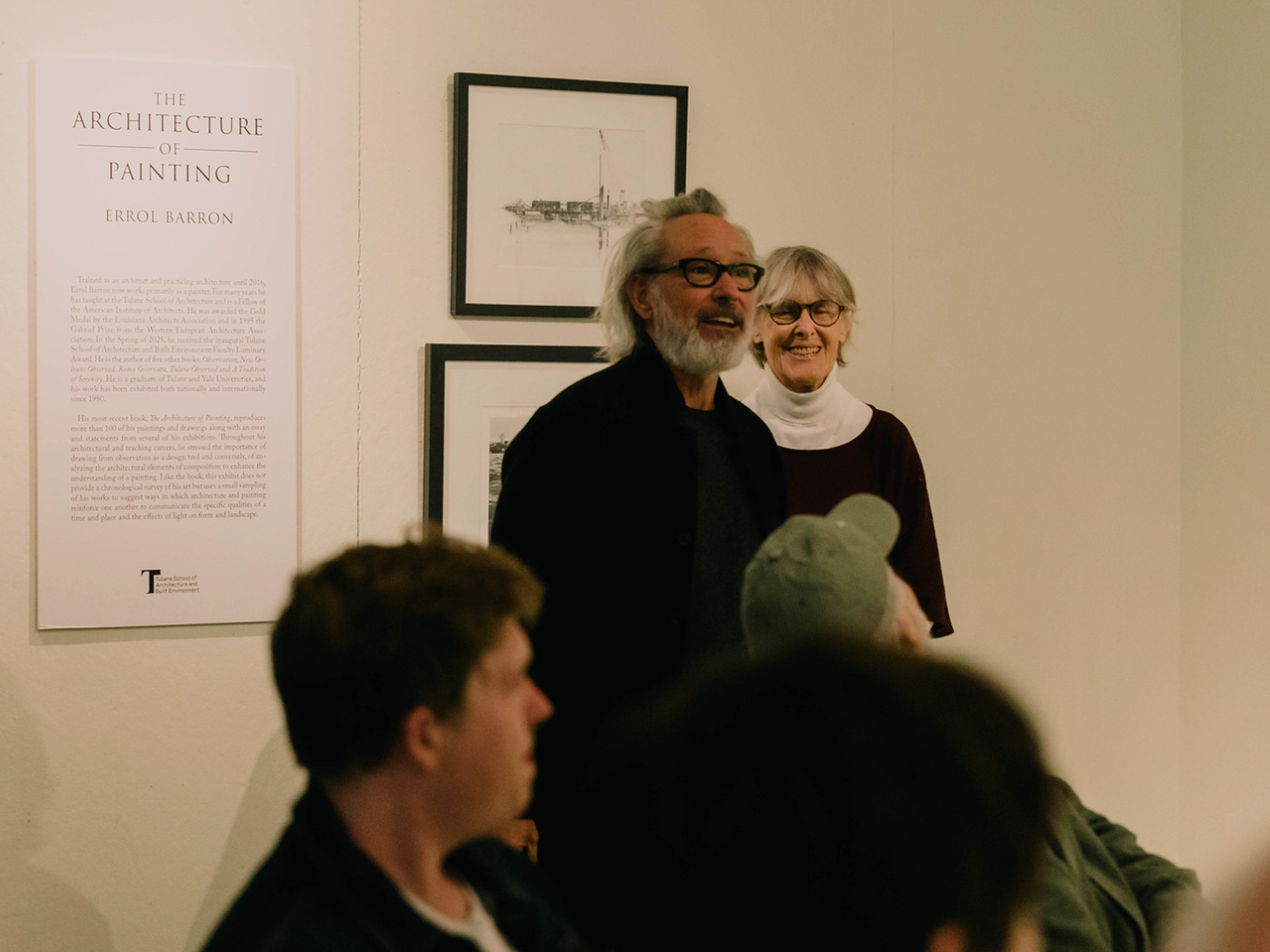 Two people standing in front of a poster reading "The Architecture of Painting, Errol Baron" with a description below