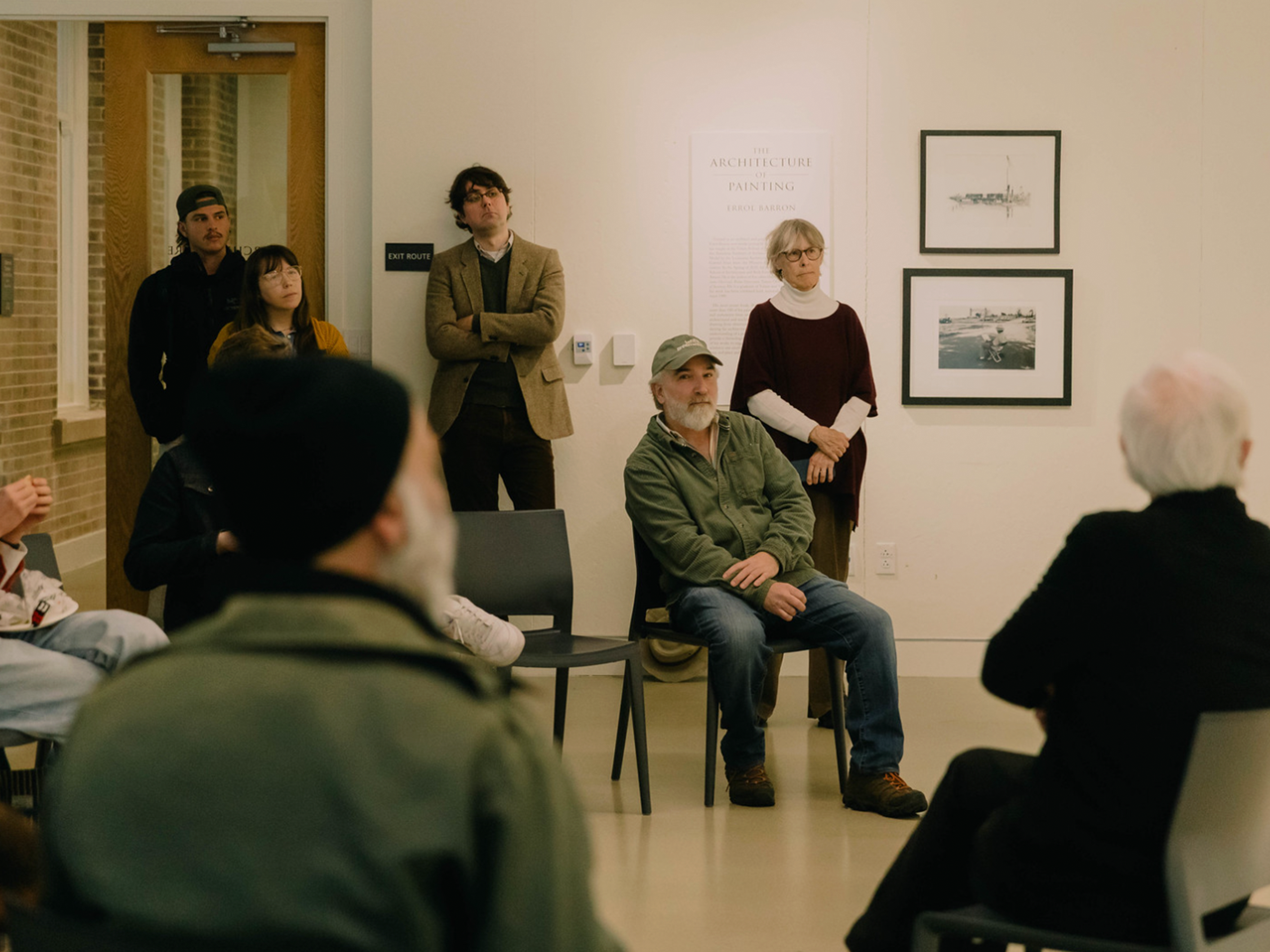 Photo of Errol Baron lecturing from behind, showing the crowd in front of him and exhibition poster