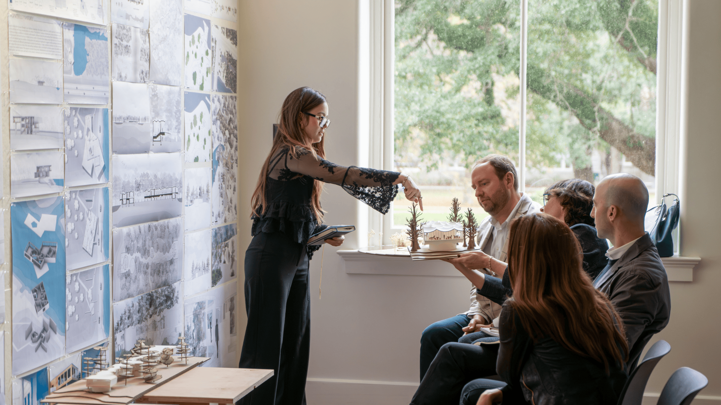 A student stands in front of a seated jury of guest reviewers, all looking at a model held by a reviewer.
