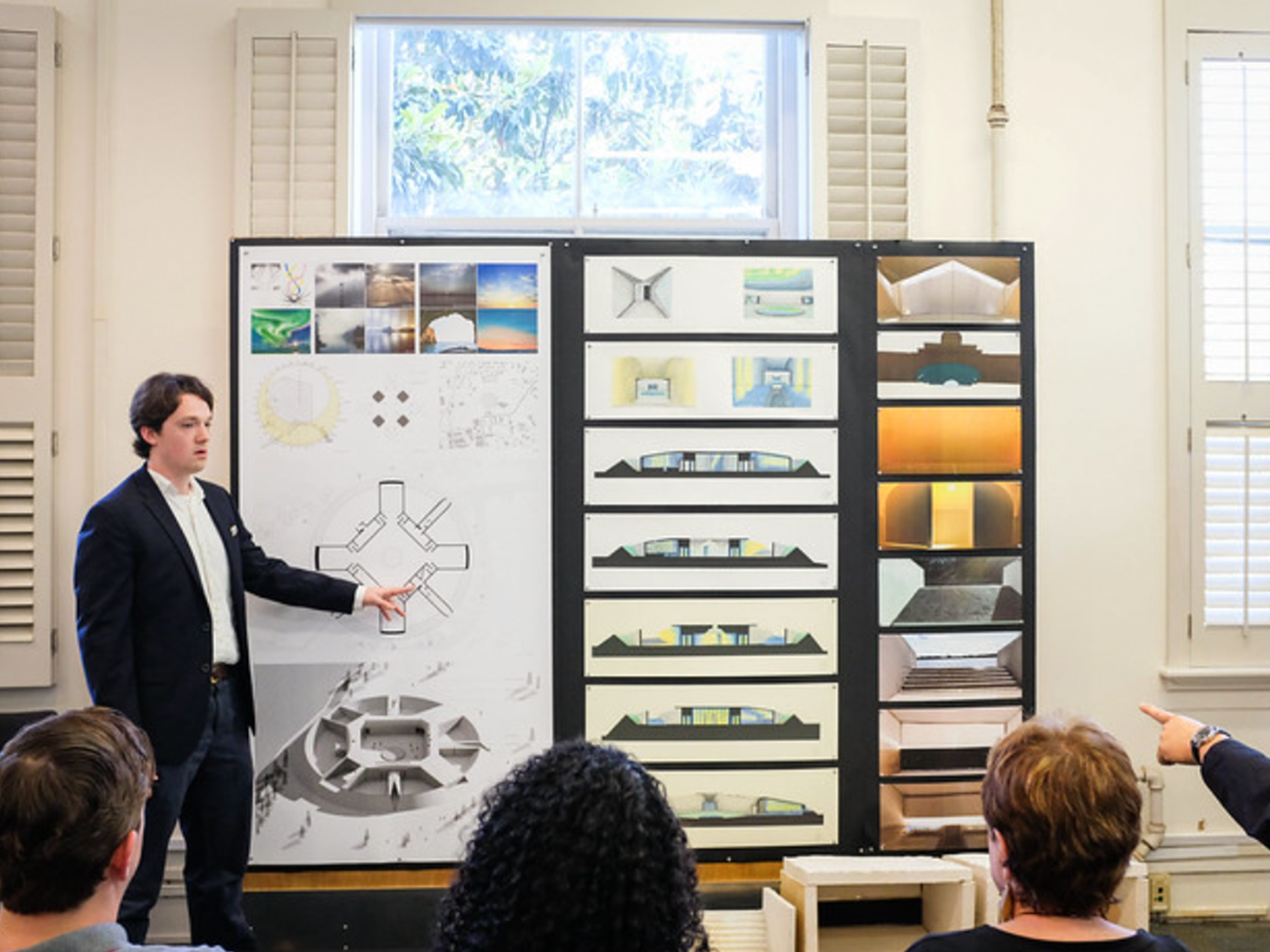 Presenter in suit standing in front of audience and pointing to rolling poster, window in the background