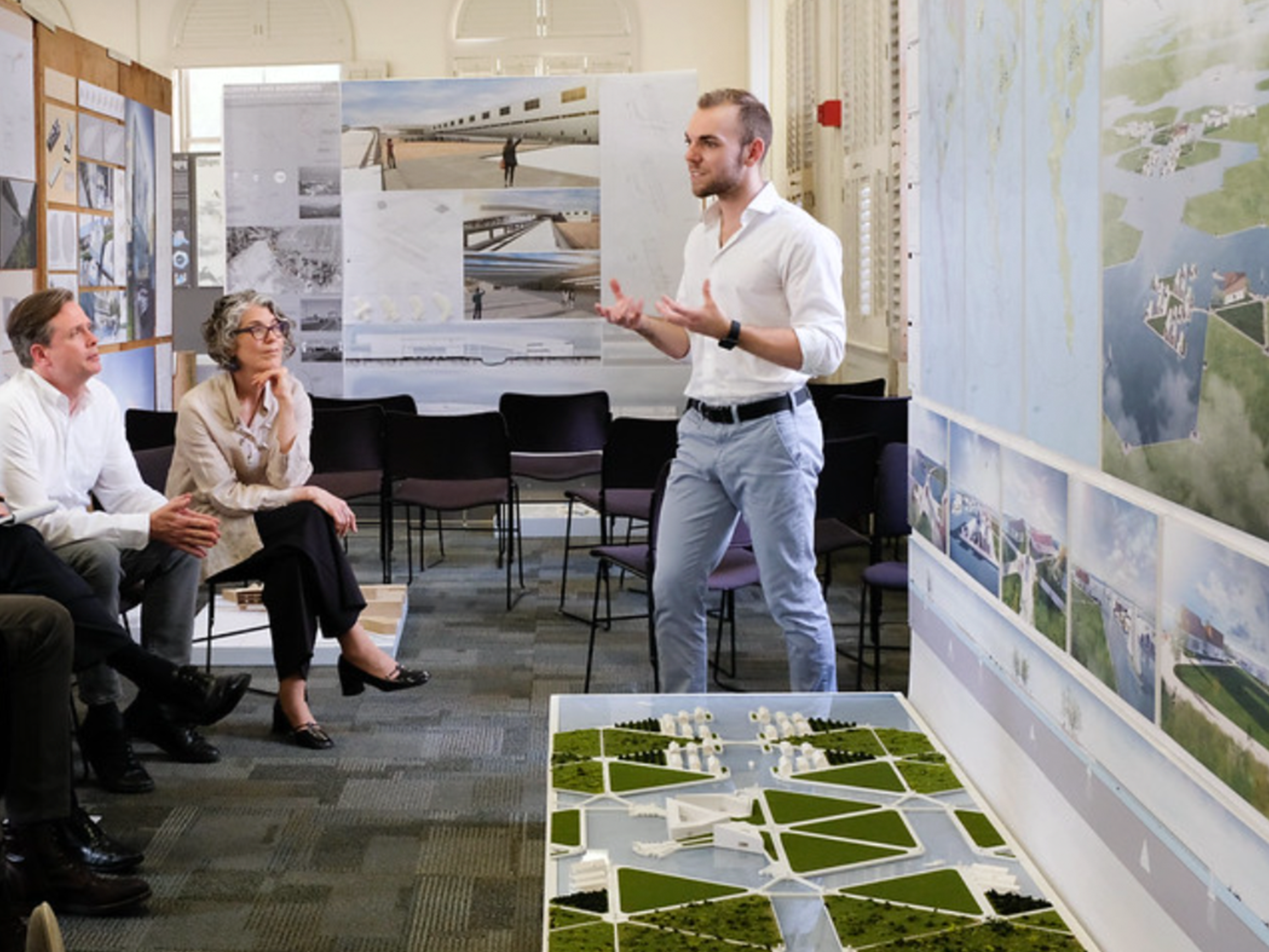 Presenter talking to the audience in front of him, presentation behind him and landscape model next to him