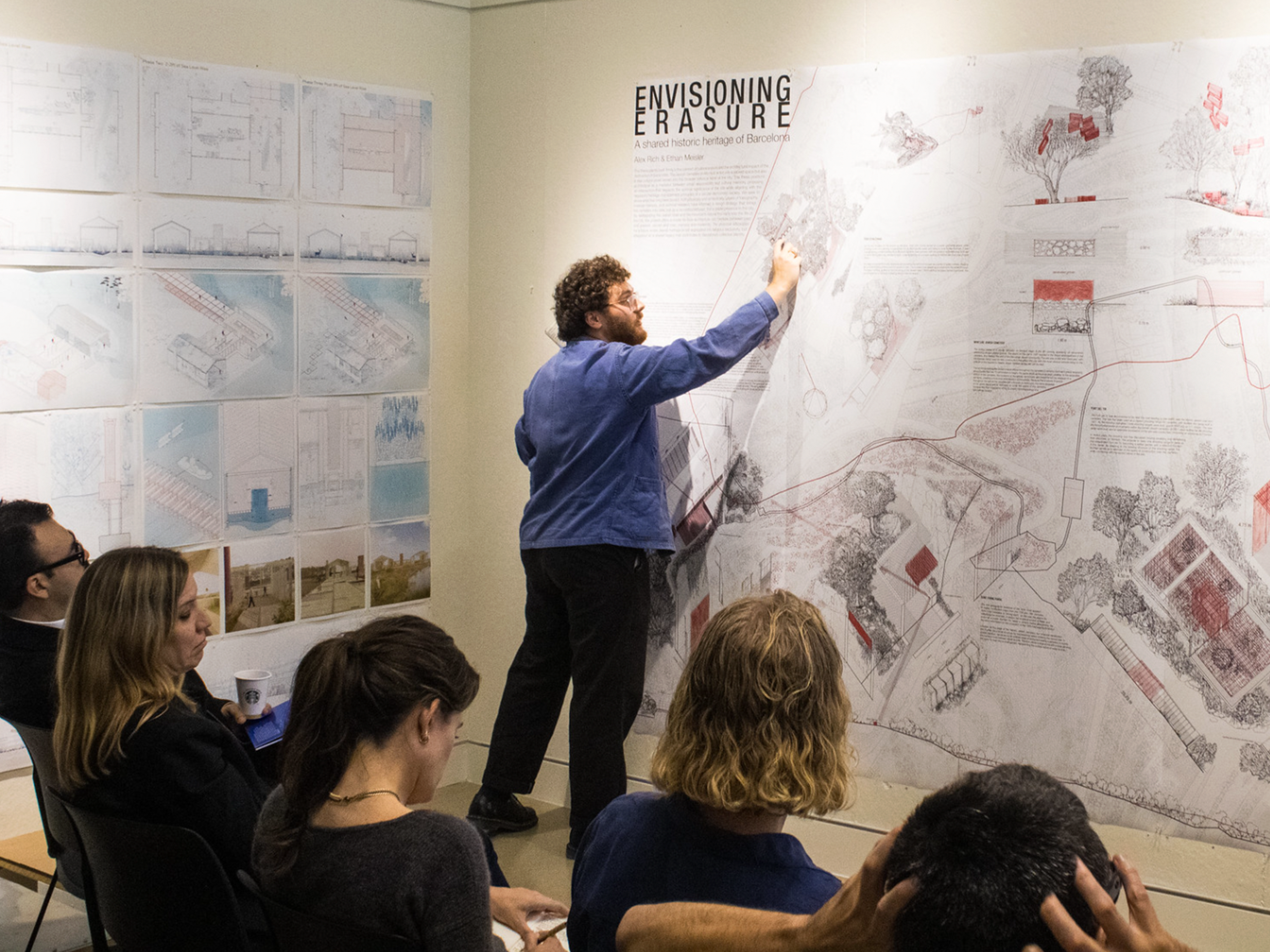 Man pointing at large printed-out plans on the wall, crowd watching