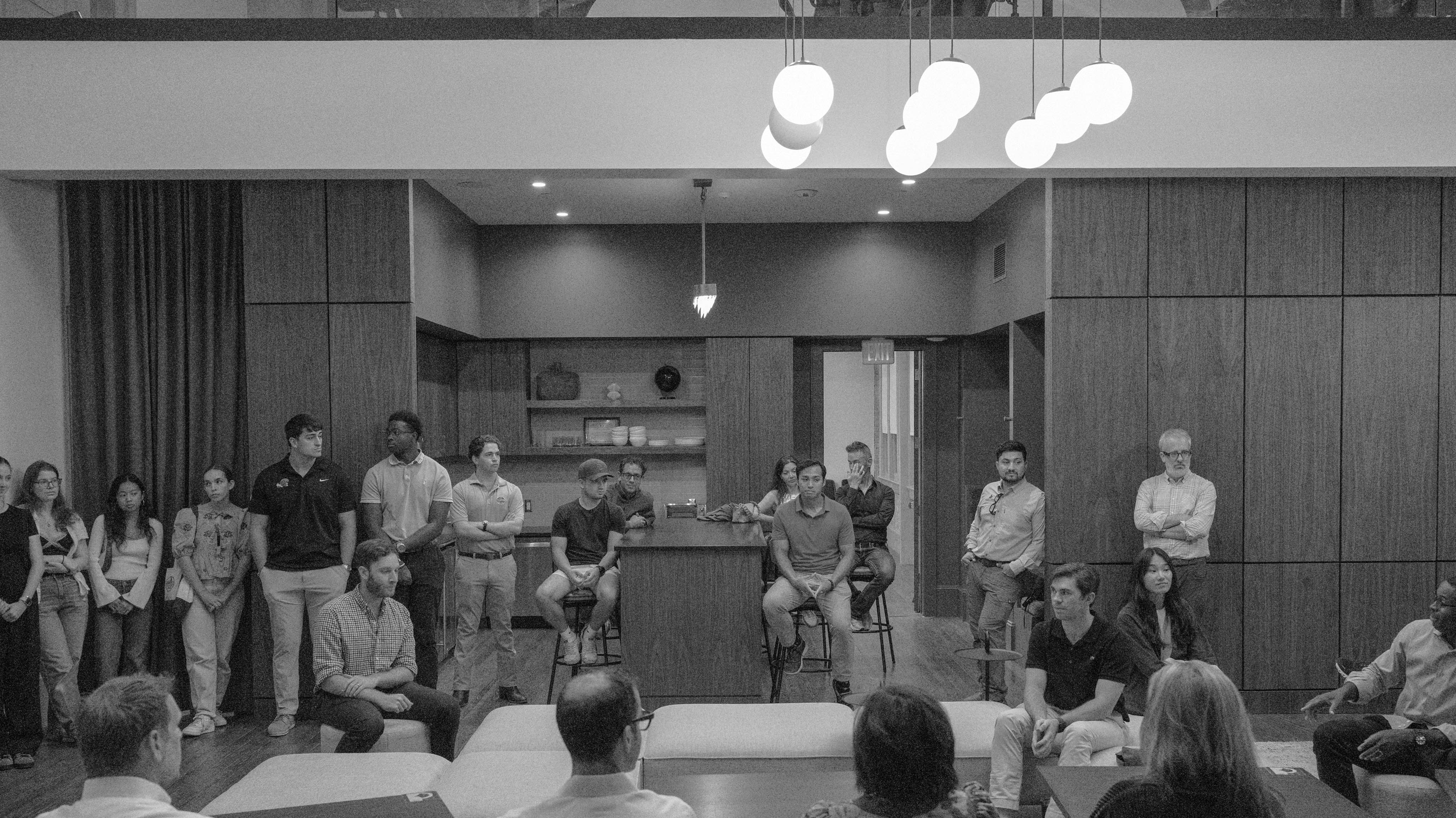 Students and faculty sit and stand while listening to a faculty talk on the right side in an open layout kitchen-dining area of a real estate development project.