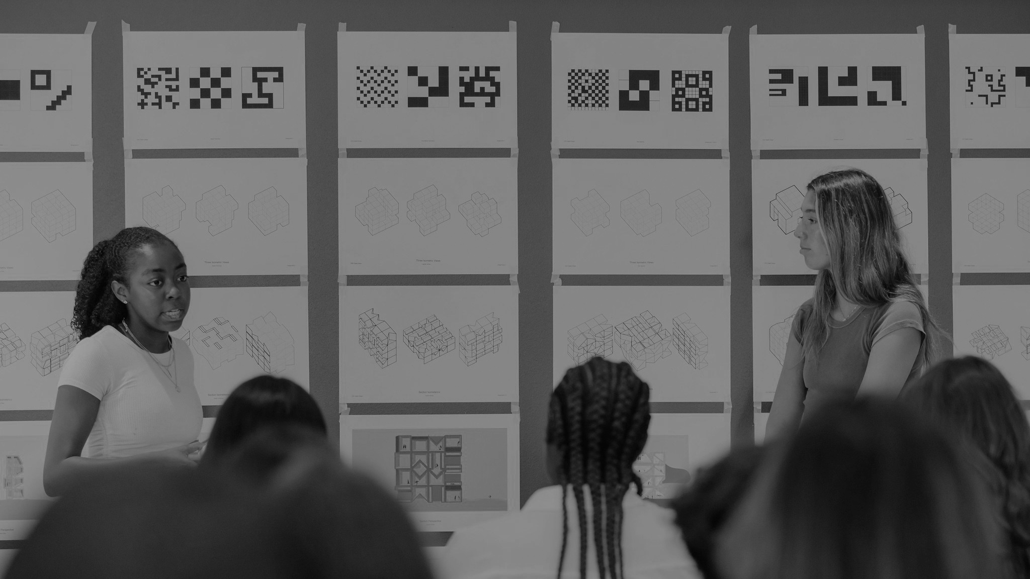 Two students presenting their work to a group of people, their work is pinned up on the wall.