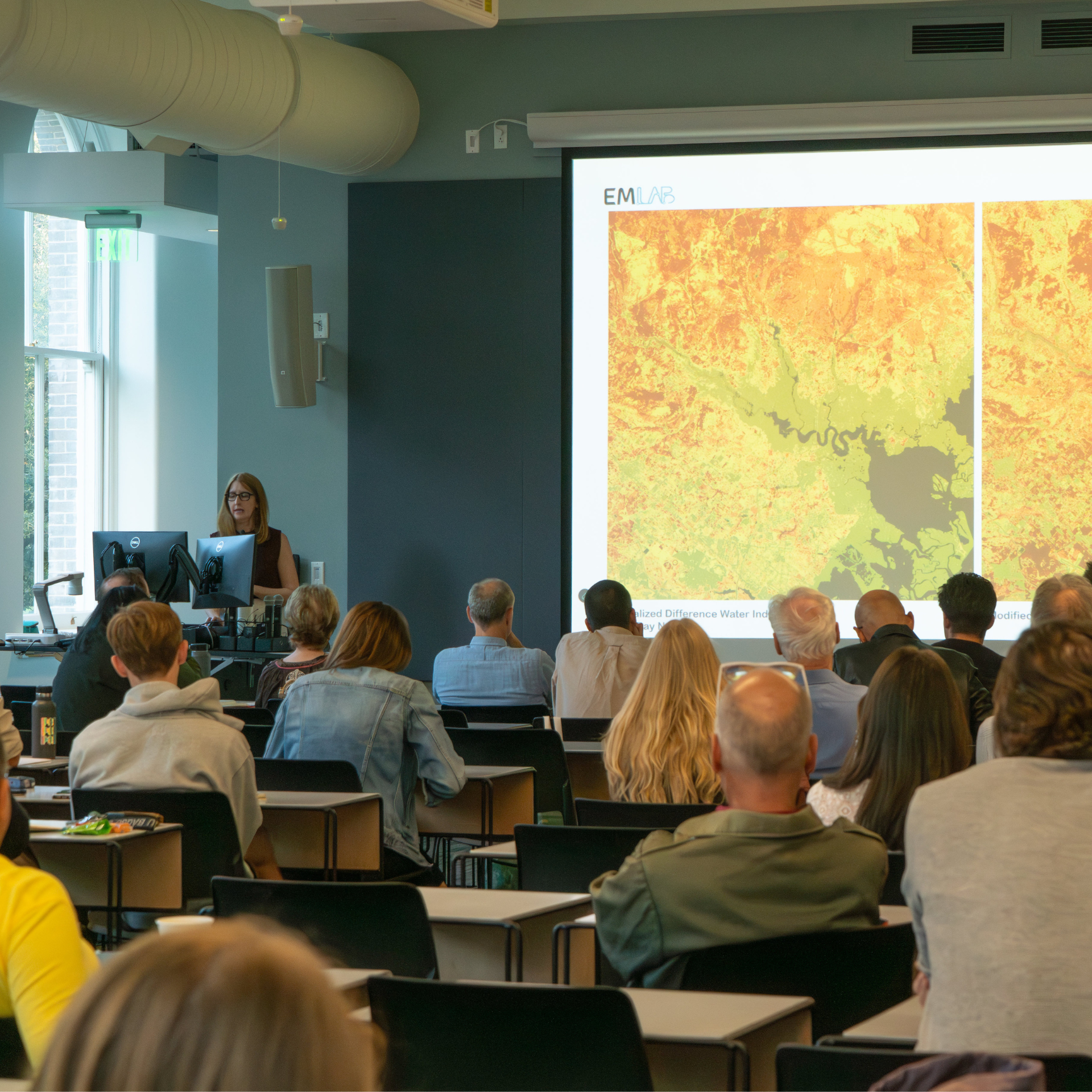 View from the back of a large lecture hall and rows of people sitting and listening to a guest lecturer in the front of the room.