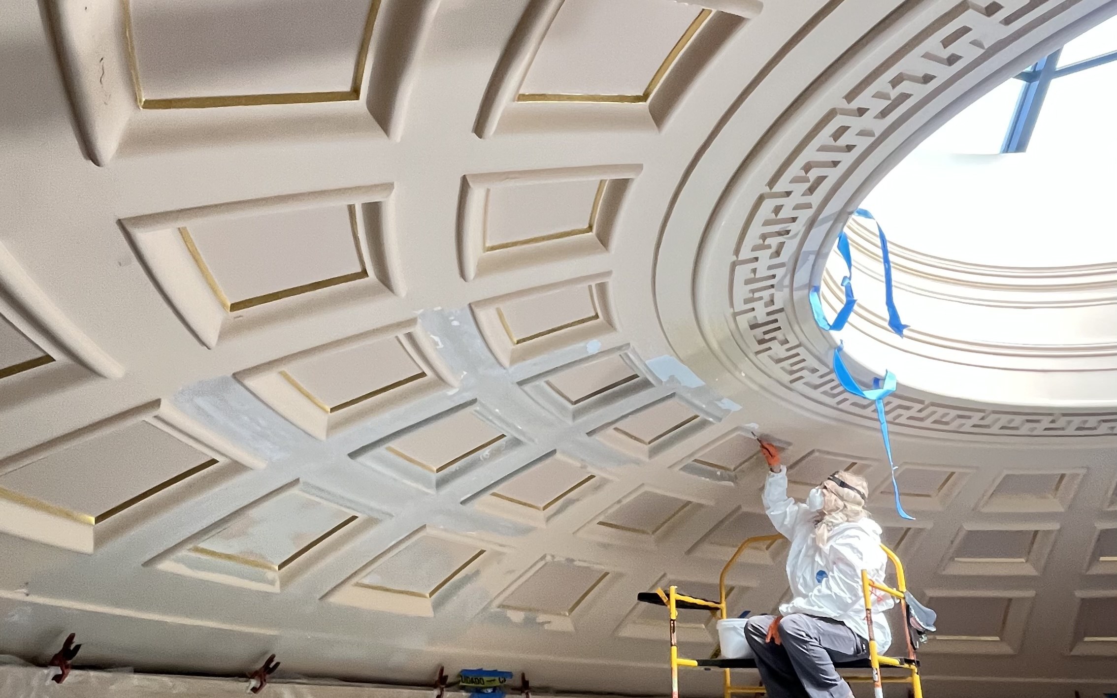 A conservationist wearing white protective gear working on the ceiling of a domed room.