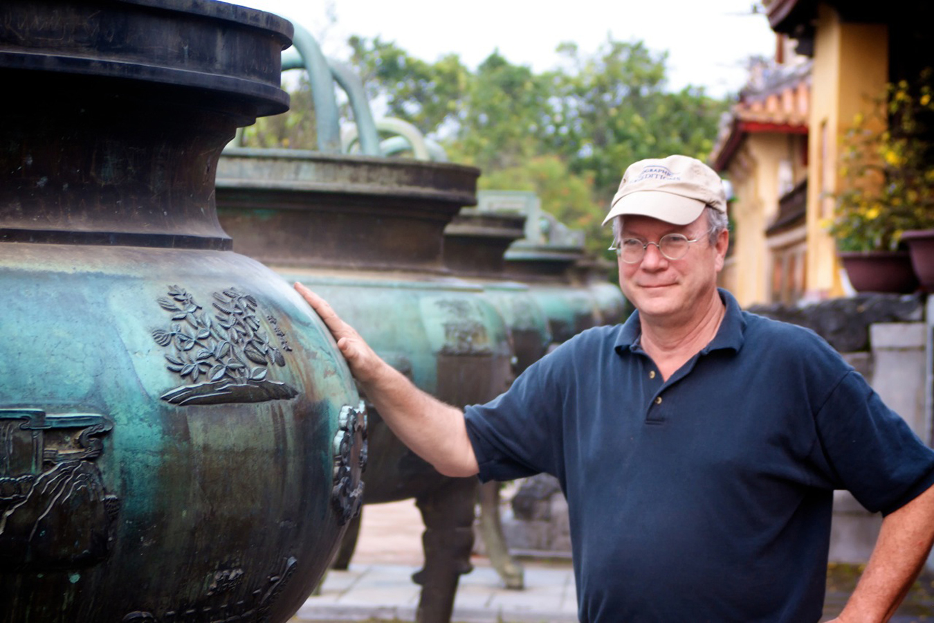 John Stubbs, wearing a navy blue collared shirt and a baseball hat, resting his arm on a sculpture to the left